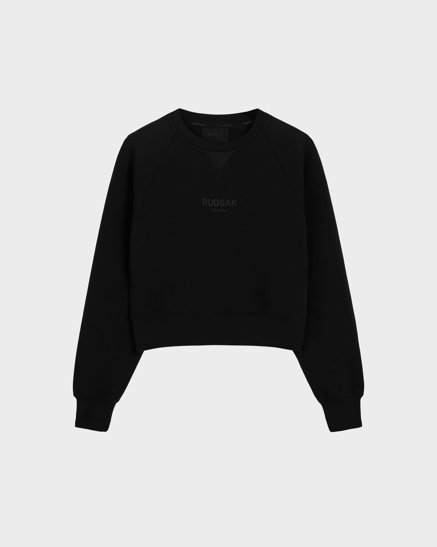 The BETSY - 8525087 Black is a black crewneck sweater by Rudsak with ribbed cuffs and hem, featuring the brand subtly printed on the front in a modern cropped silhouette, shown against a plain light grey background.