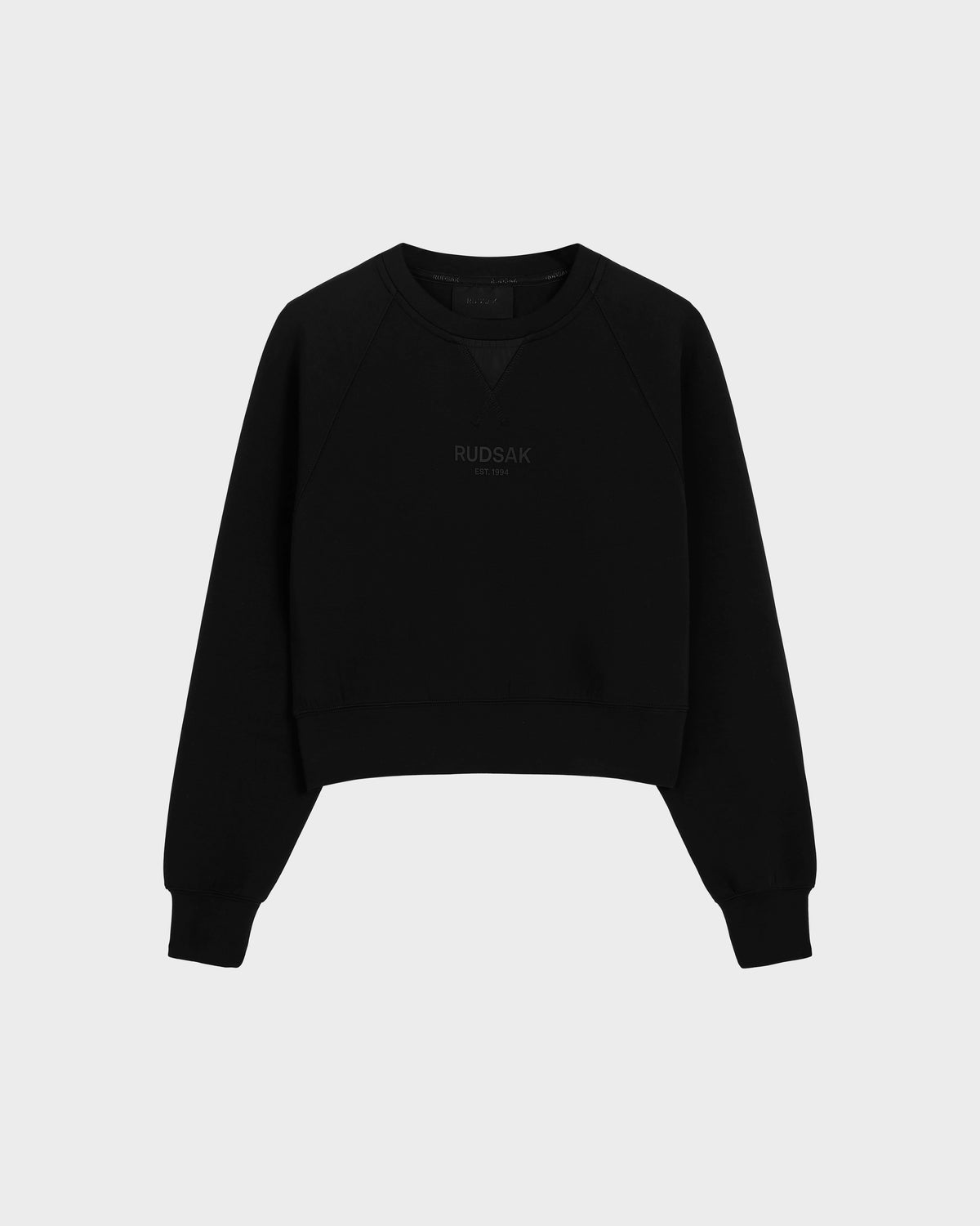 The BETSY - 8525087 Black is a black crewneck sweater by Rudsak with ribbed cuffs and hem, featuring the brand subtly printed on the front in a modern cropped silhouette, shown against a plain light grey background.