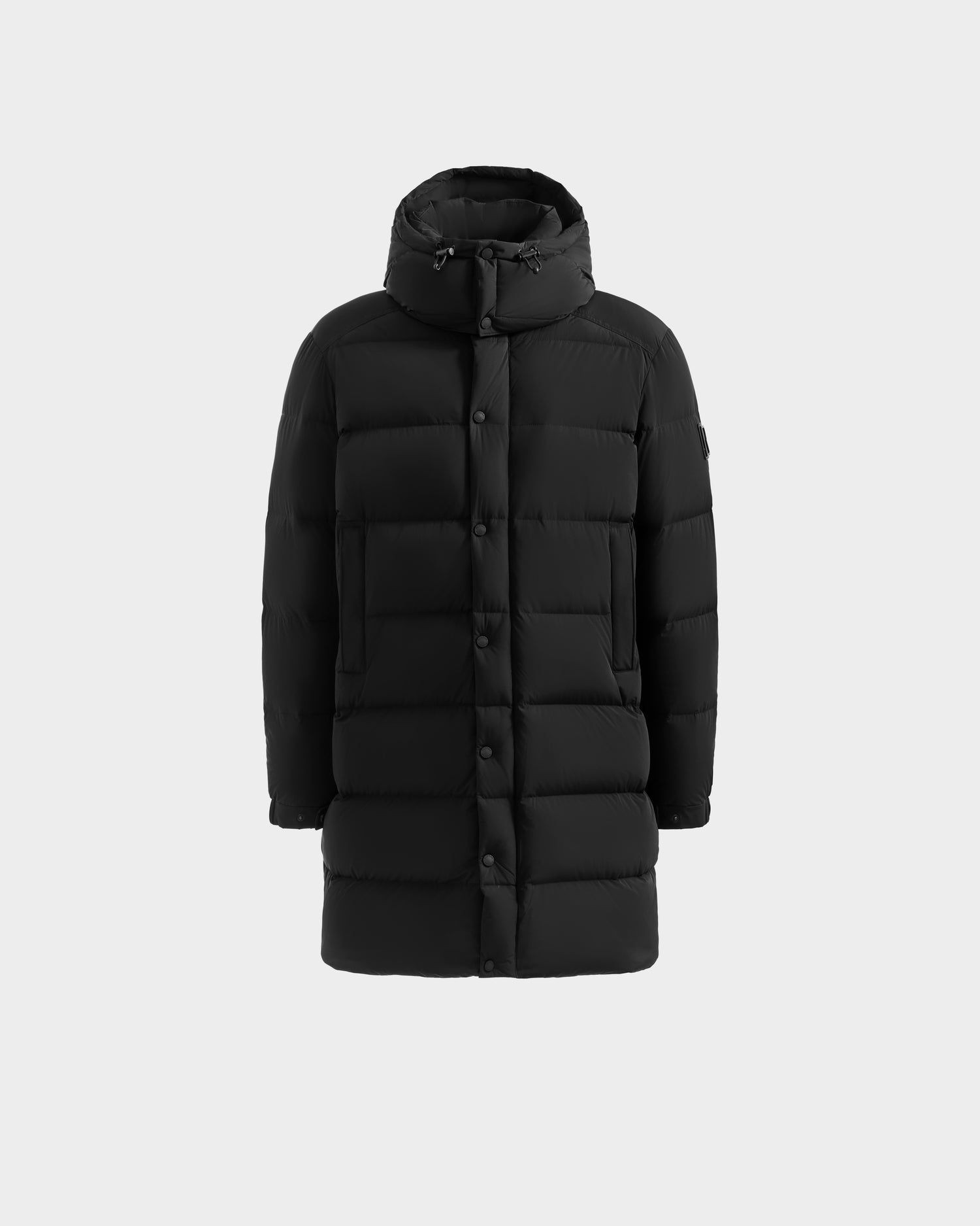 The Rudsak BRAYDEN M-M - M125535 Black is a men’s long down puffer jacket with a hood, front snap buttons, and horizontal quilting, shown against a light gray background.