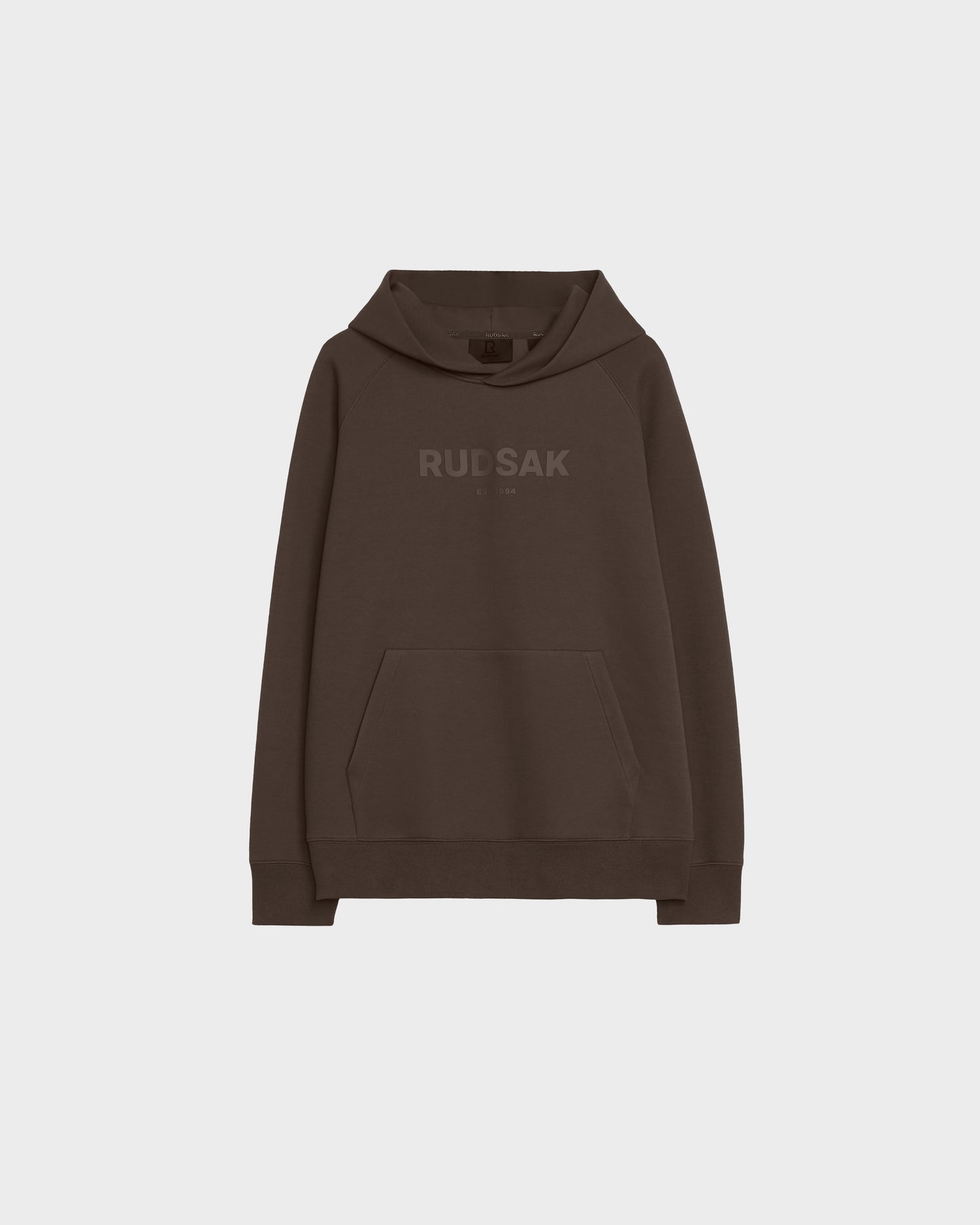 The Rudsak BRIAR - 9525001 Espresso W is a dark brown unisex hoodie with a front kangaroo pocket and RUDSAK in matching brown on the chest, shown on a plain light gray background.