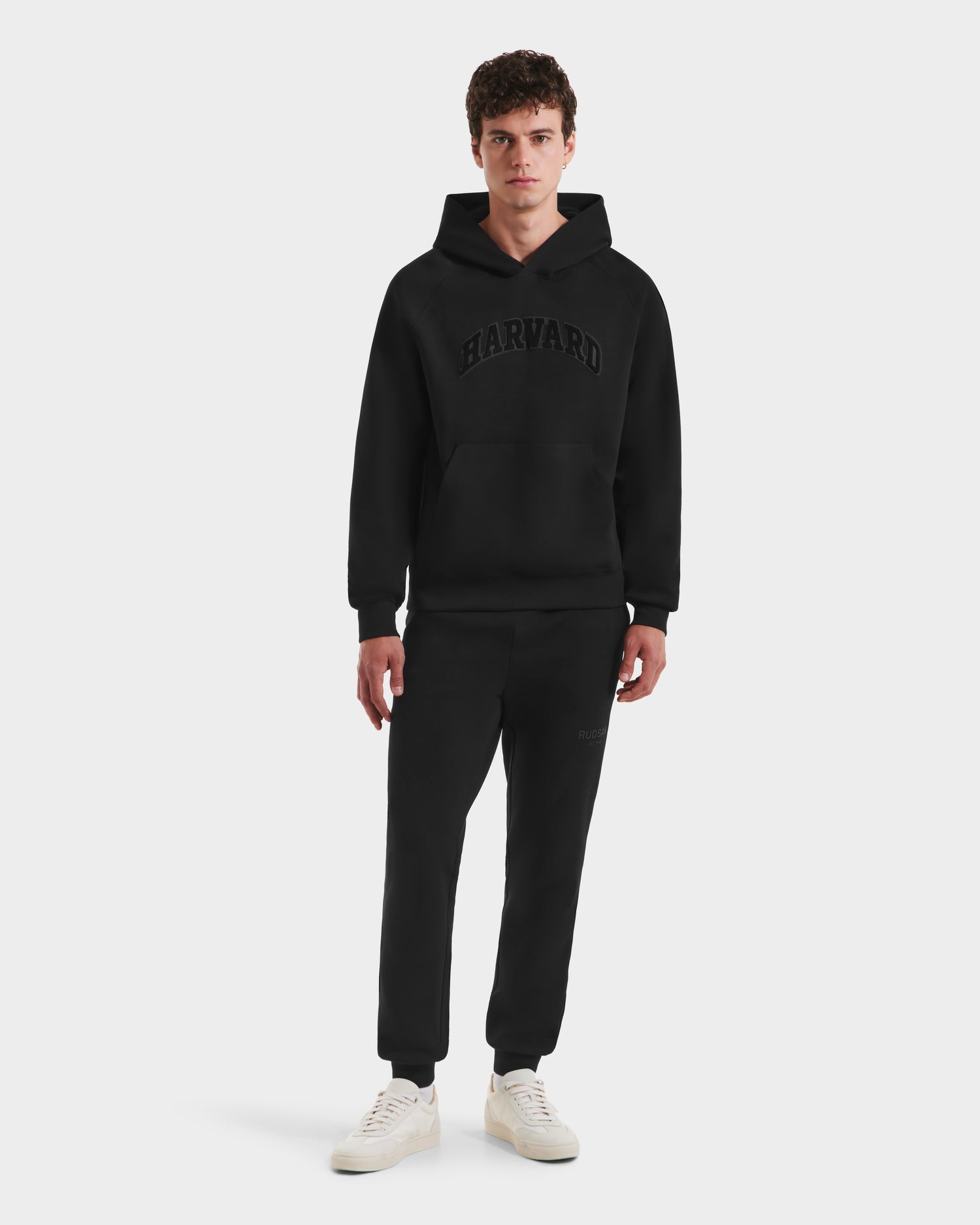 A young man faces forward in a limited-edition Rudsak BRIAR B HV - H525509 black hoodie and matching sweatpants, paired with white sneakers. The setting is a plain light gray background.