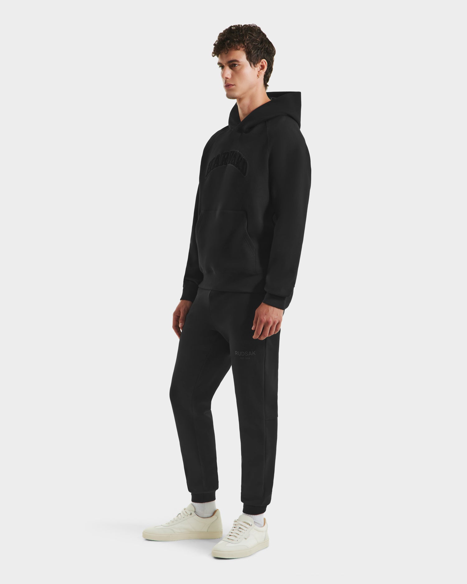 A person stands against a white background wearing the Rudsak BRIAR B HV - H525509 Black hoodie with a front pocket and subtle chest text, paired with black joggers and white sneakers.