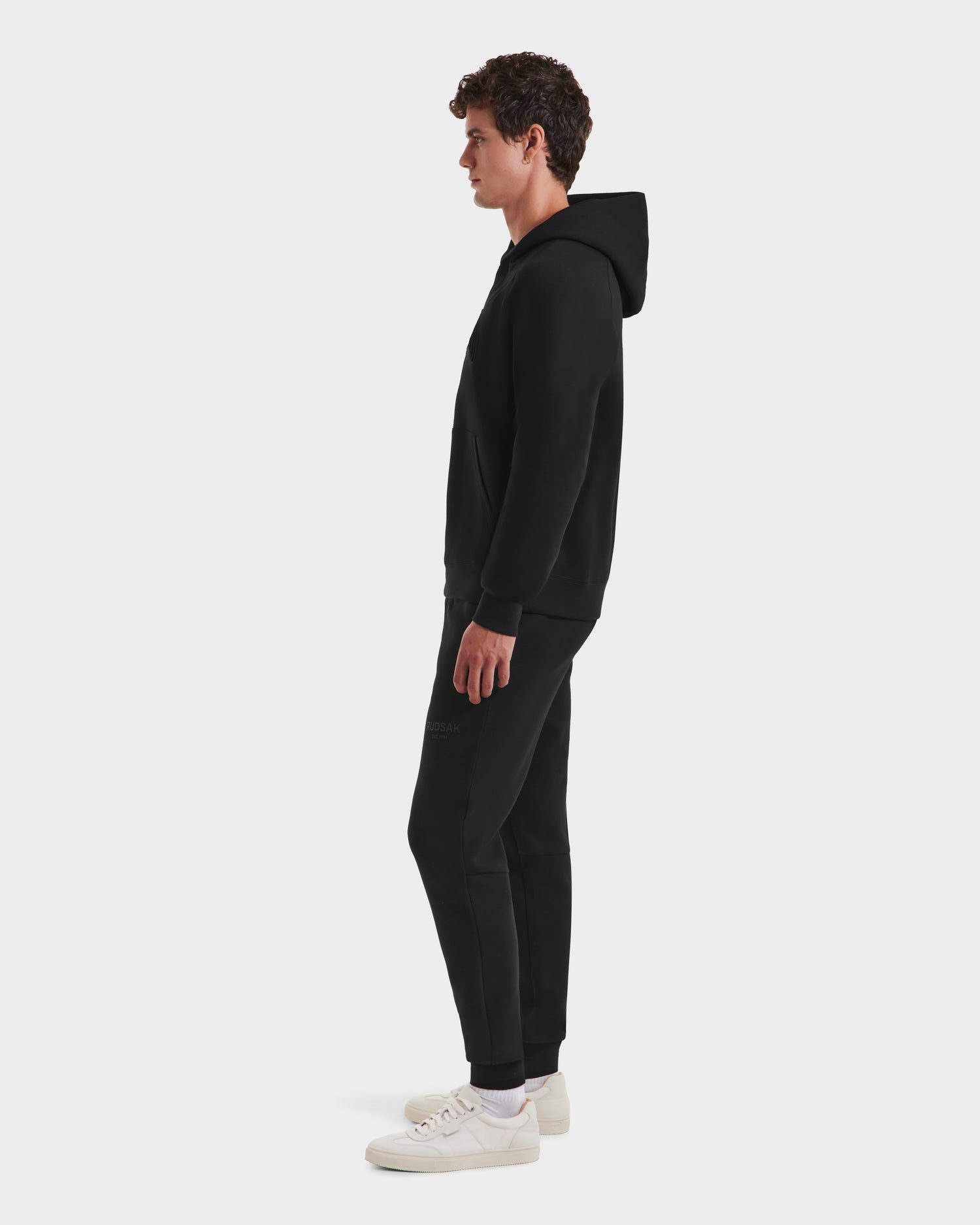 A man in profile wears the limited-edition Rudsak BRIAR B HV - H525509 black hoodie and matching joggers with white sneakers, standing against a plain white background.