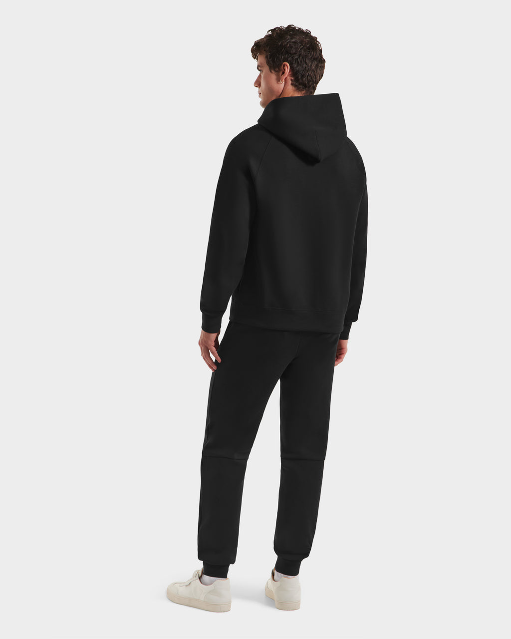 A person stands facing away in a limited-edition Harvard hoodie, Rudsak BRIAR B HV - H525509 Black joggers, and white sneakers against a plain light background.