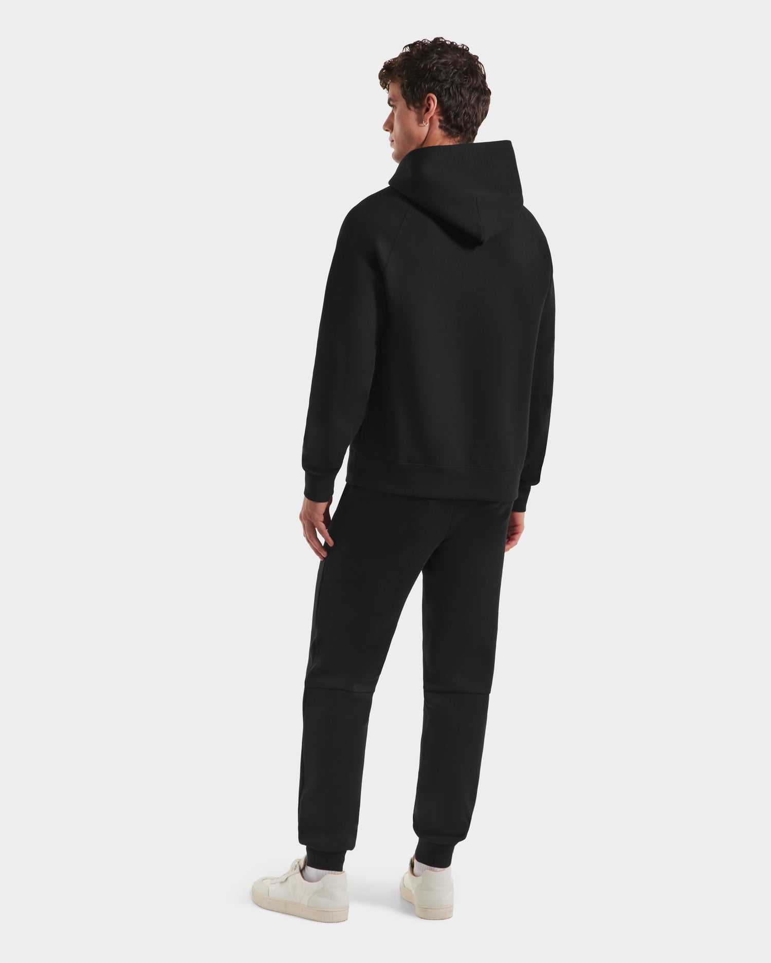 A person stands facing away in a limited-edition Harvard hoodie, Rudsak BRIAR B HV - H525509 Black joggers, and white sneakers against a plain light background.