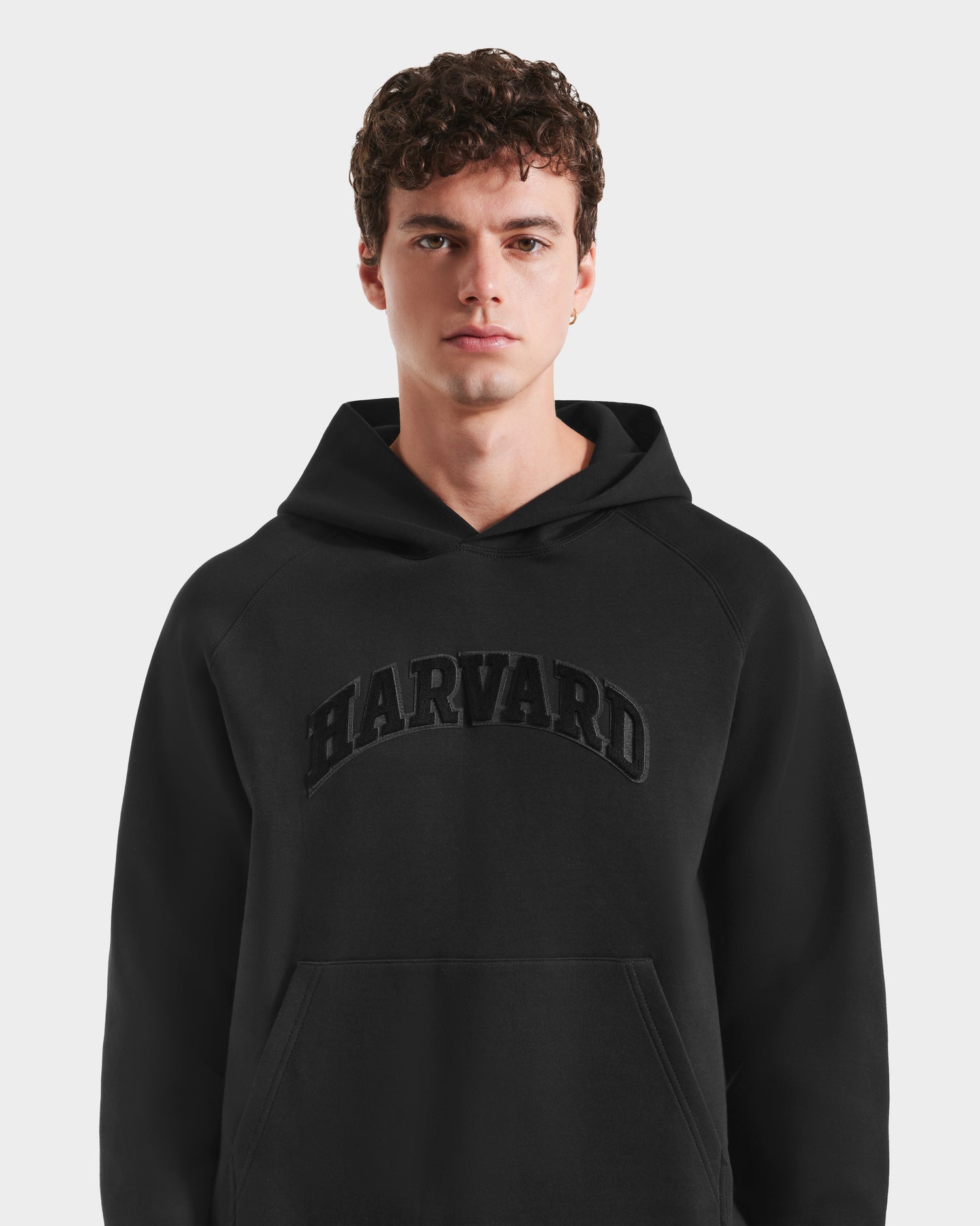 A young man with curly brown hair models the Rudsak BRIAR B HV - H525509 Black hoodie, its striking design highlighted as he poses against a plain white background.