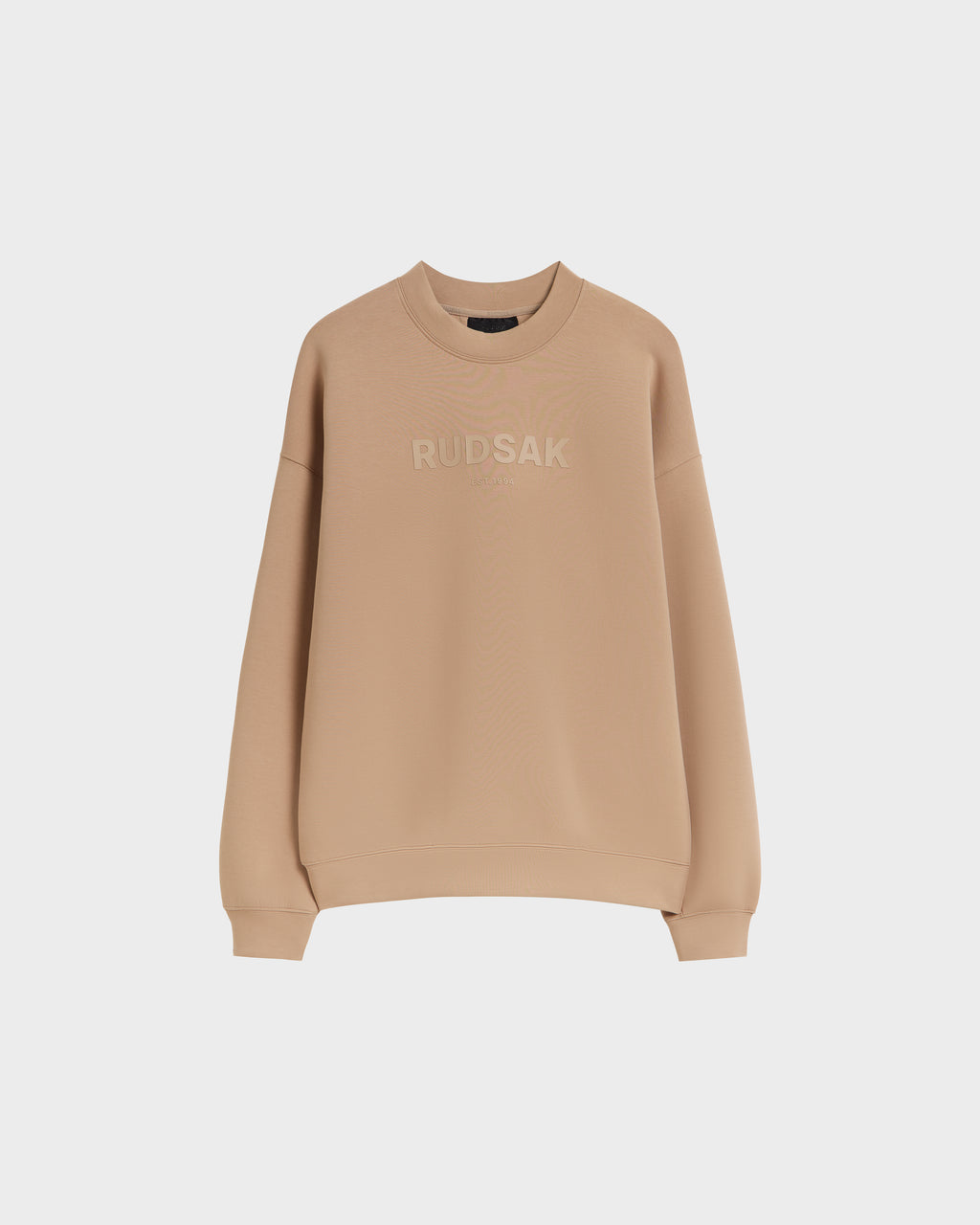 The Rudsak BRYN - 9524007 Almond M is a beige cotton crewneck sweatshirt with long sleeves, ribbed cuffs and hem, and tonal “RUDSAK” chest embroidery. Genderless fit for everyday comfort, shown here against a white background.