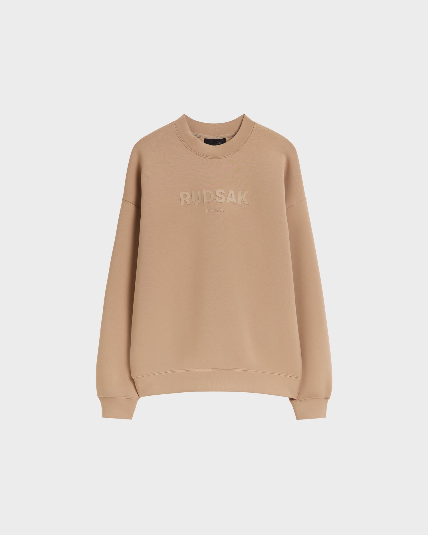 The Rudsak BRYN - 9524007 Almond W is a beige cotton crewneck sweatshirt with matching beige RUDSAK embroidery on the chest, long sleeves, and genderless sizing, shown on a plain white background.