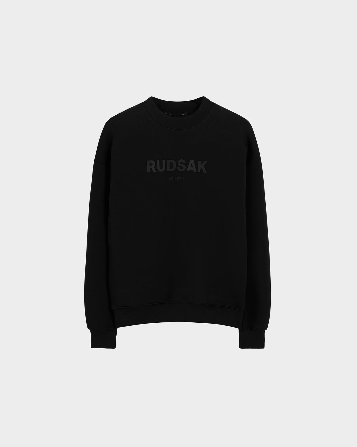 The BRYN - 9524007 Black W crewneck sweatshirt by Rudsak features subtle dark RUDSAK and EST. 1994 prints on the front, crafted from black cotton with genderless sizing—displayed on a light grey background for versatile everyday style.