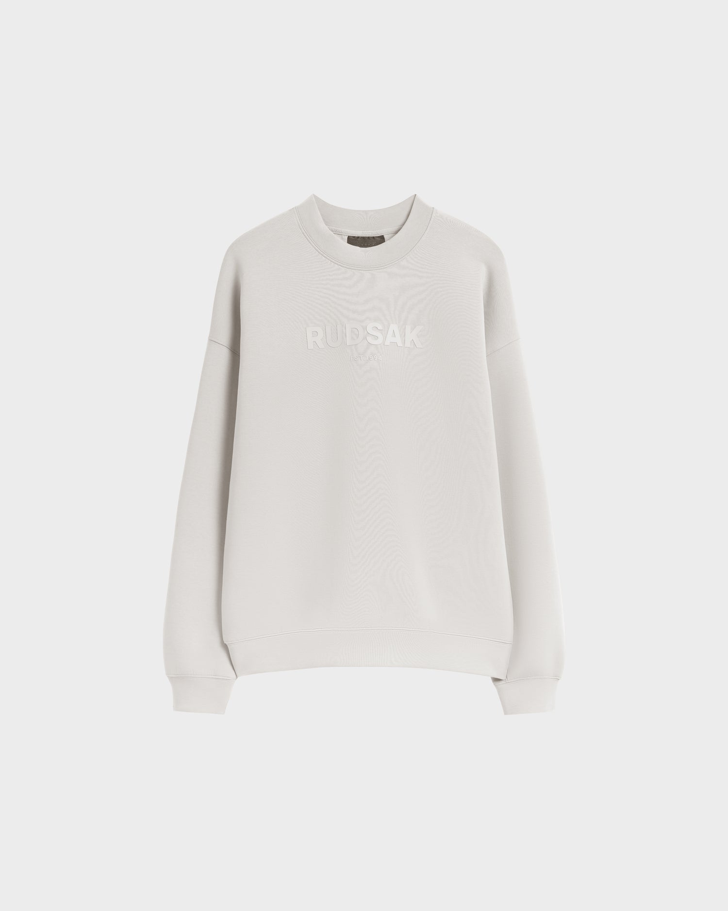 The Rudsak BRYN - 9524007 Vanilla M is a light grey, cotton crewneck sweatshirt with long sleeves and tonal RUDSAK embroidery on the chest. It offers genderless sizing for a comfortable everyday fit, shown on a white background.