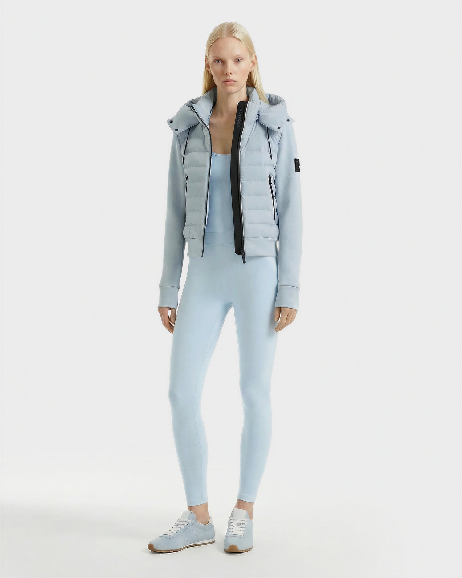A woman faces forward wearing the Rudsak CASSA - 8125057 Powder Blue fitted bodysuit, styled with a matching puffer vest jacket featuring a black zipper and coordinating sneakers, set against a plain white background.