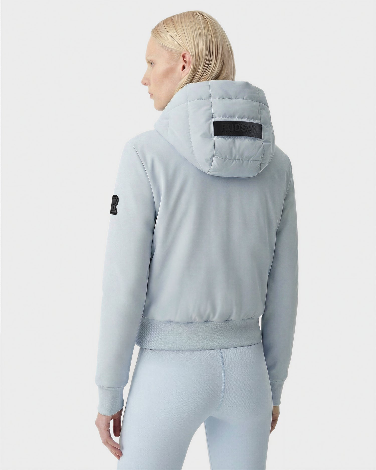 A person with straight, light blonde hair wears the Rudsak CASSA - 8125057 Powder Blue women's transitional jacket and matching pants, featuring a patch and visible logo on the hood. The background is plain white.