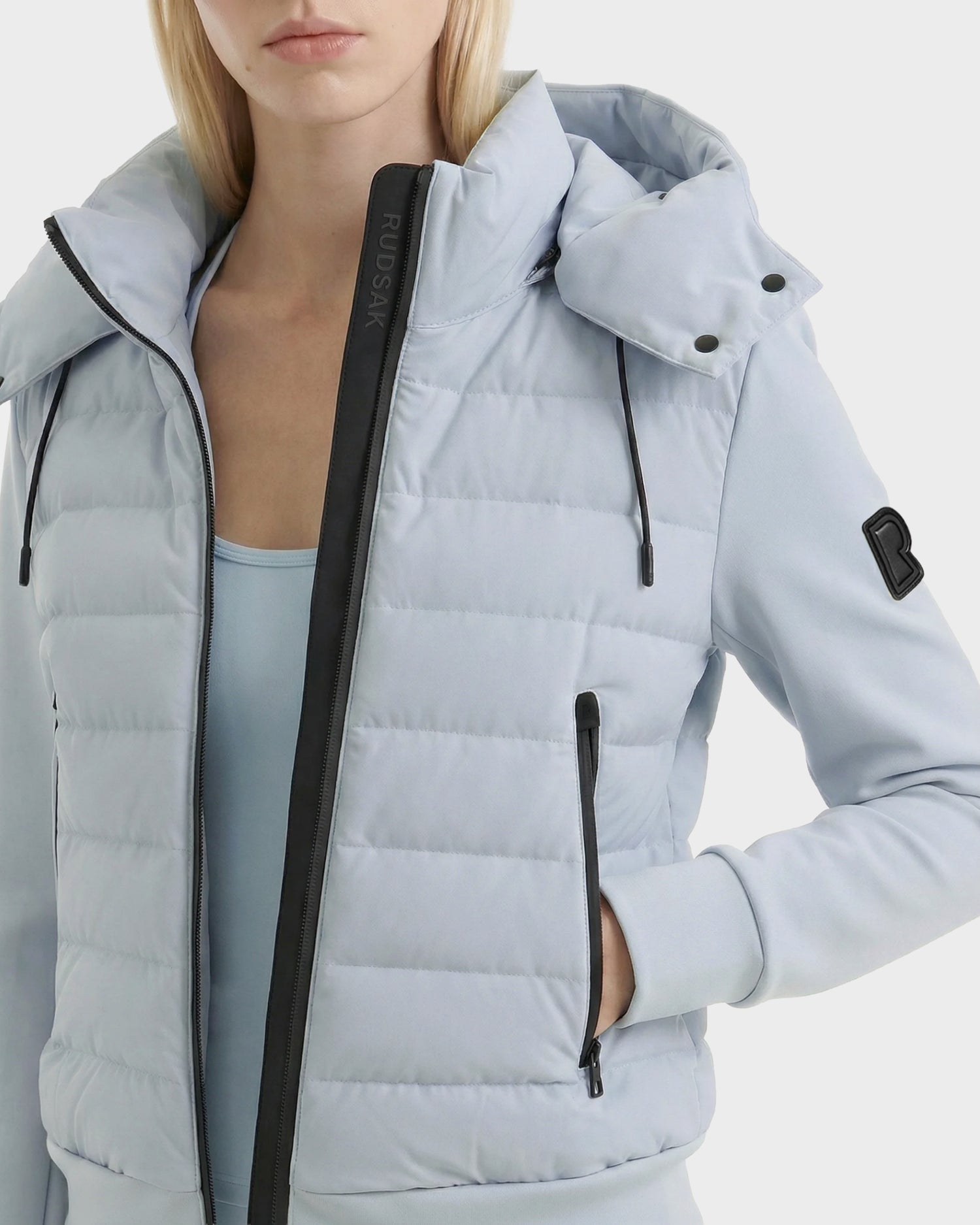 A woman wears the Rudsak CASSA - 8125057 Powder Blue transitional jacket with black zippers and a sleeve patch, standing against a white background with one hand in her pocket.
