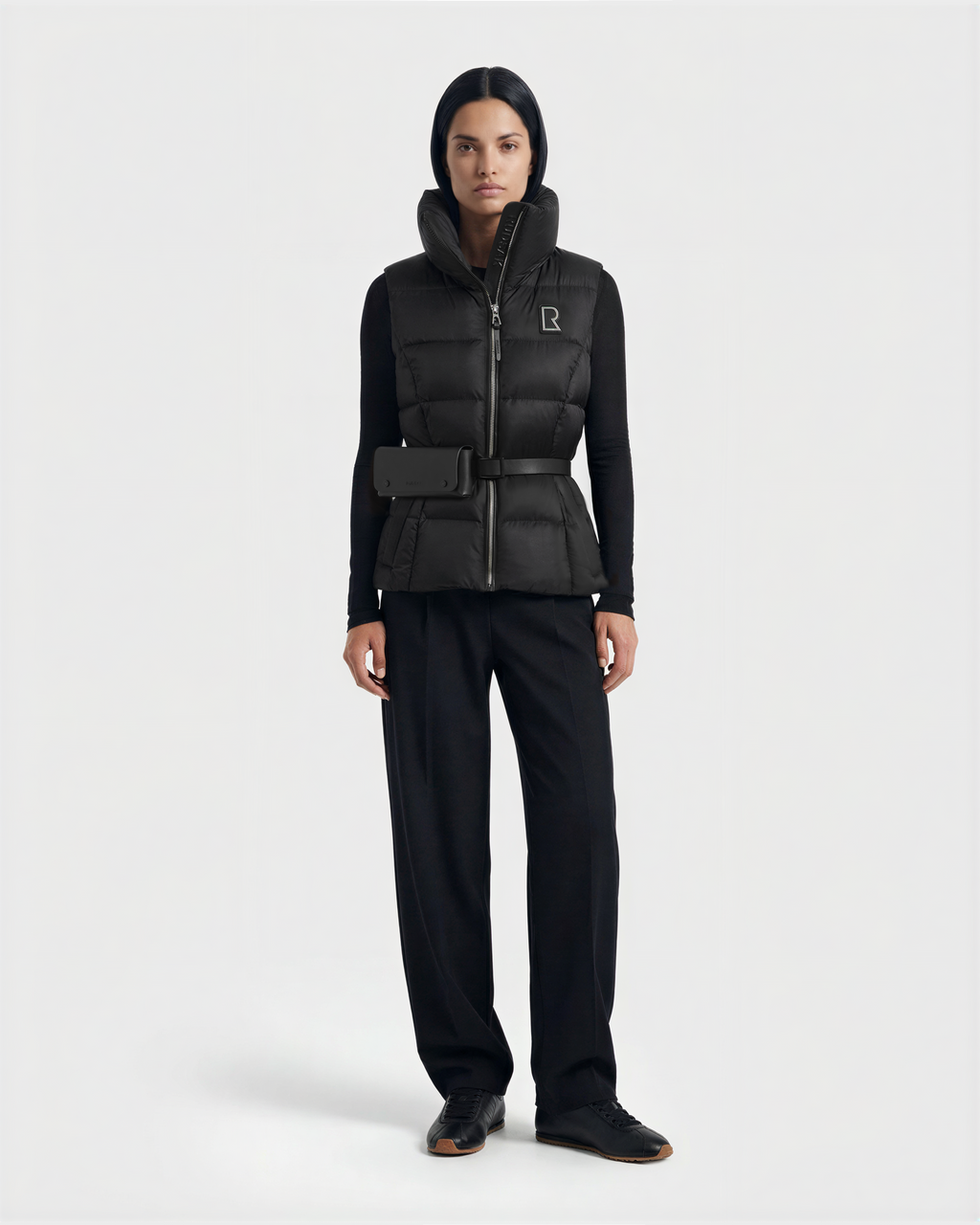 A woman faces forward against a plain light background, wearing the Rudsak CASSIDY N - 8125677 Black women’s down vest with a high collar, paired with a black long-sleeve top, black trousers, and black shoes.