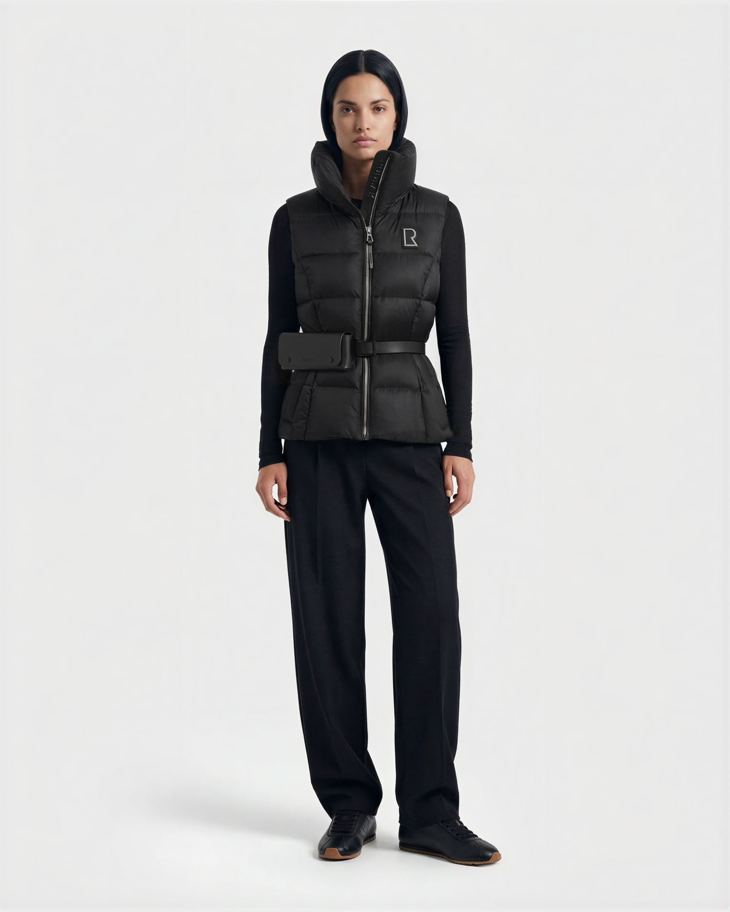 A woman faces forward against a plain light background, wearing the Rudsak CASSIDY N - 8125677 Black women’s down vest with a high collar, paired with a black long-sleeve top, black trousers, and black shoes.
