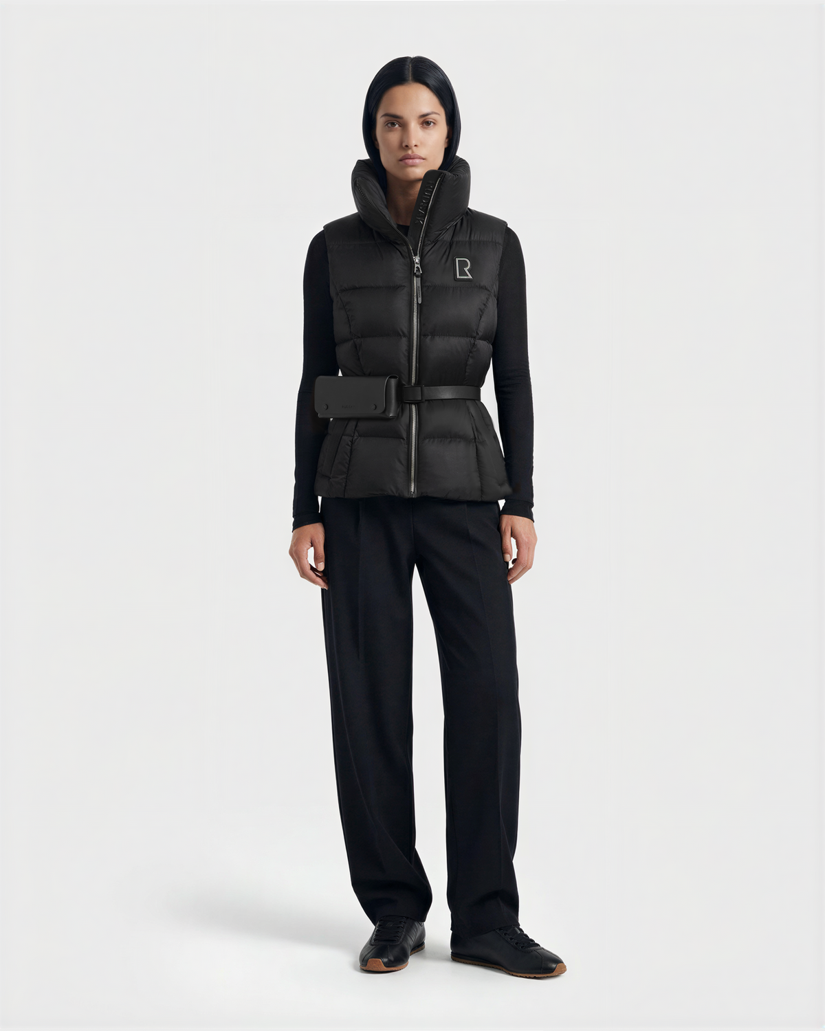 A woman faces forward against a plain light background, wearing the Rudsak CASSIDY N - 8125677 Black women’s down vest with a high collar, paired with a black long-sleeve top, black trousers, and black shoes.