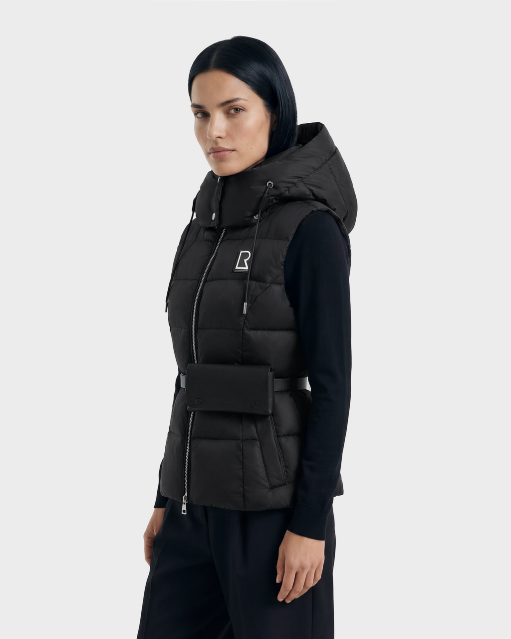 A woman with straight black hair wears the Rudsak CASSIDY N - 8125677 Black fitted down vest over a black long-sleeve top and pants, accessorized with a small black belt bag, standing against a light gray background.