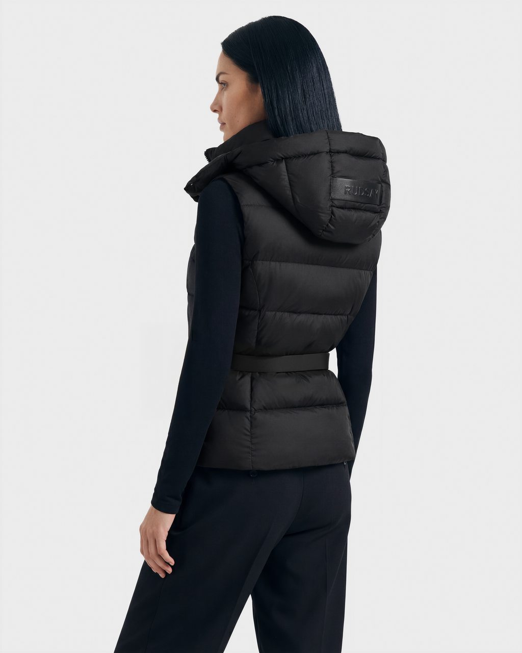 A woman with straight dark hair is seen from the back, modeling the Rudsak CASSIDY N - 8125677 Black fitted down vest over a long-sleeve black top and pants against a plain light background for lightweight warmth and modern style.