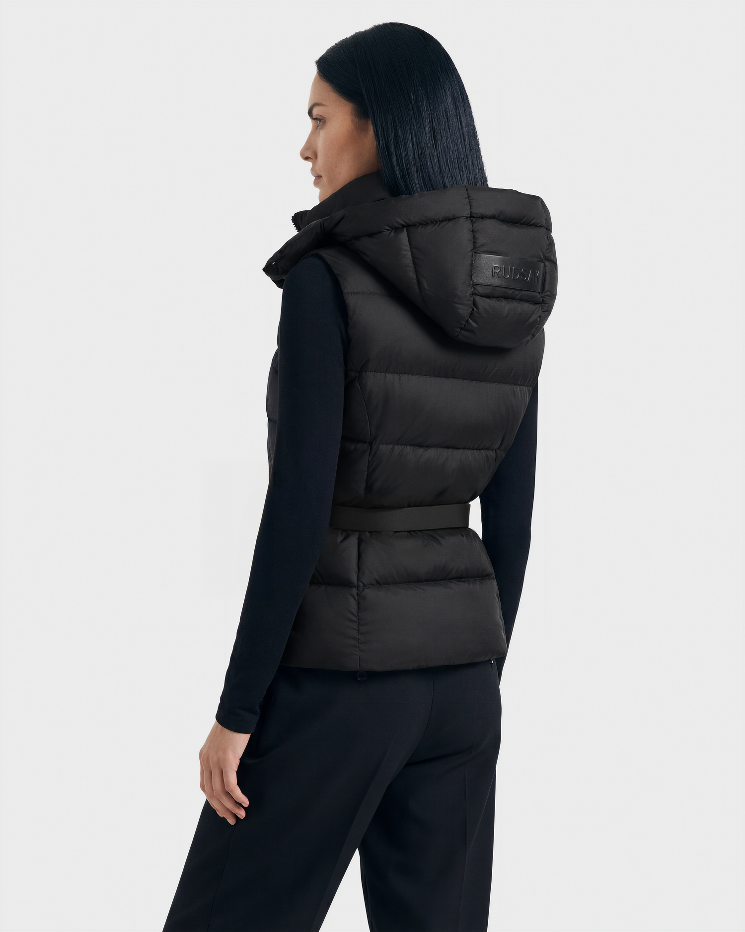 A woman with straight dark hair is seen from the back, modeling the Rudsak CASSIDY N - 8125677 Black fitted down vest over a long-sleeve black top and pants against a plain light background for lightweight warmth and modern style.