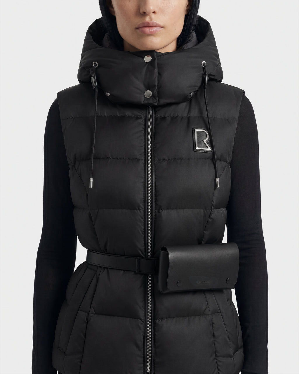 A woman wears the Rudsak CASSIDY N - 8125677 Black down vest, featuring a hood, belted waist with pouch, lightweight warmth, zipper, snap buttons, and an embroidered R logo on the left chest.