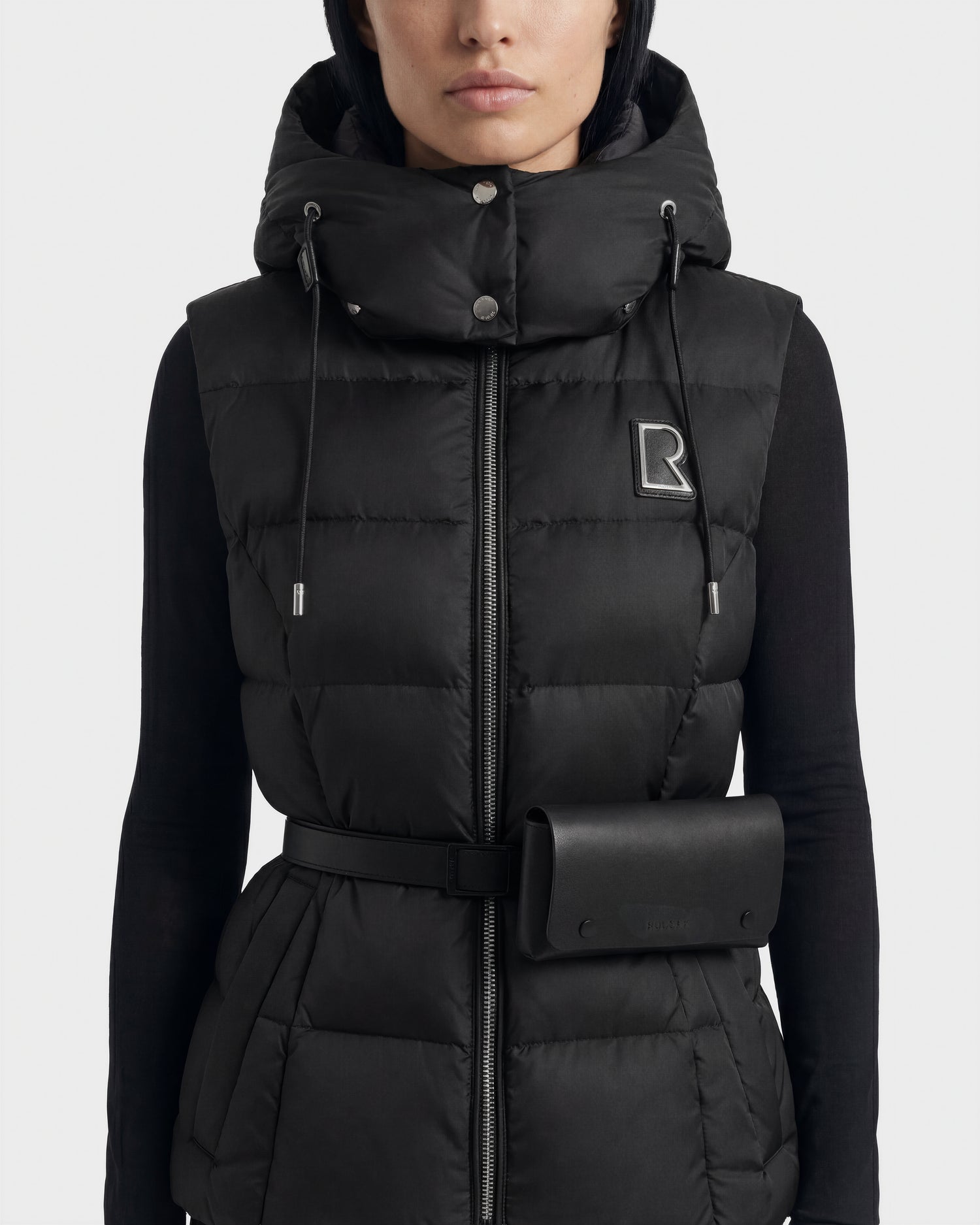 A woman wears the Rudsak CASSIDY N - 8125677 Black down vest, featuring a hood, belted waist with pouch, lightweight warmth, zipper, snap buttons, and an embroidered R logo on the left chest.