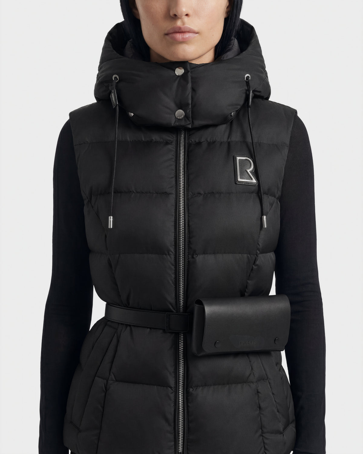 A woman wears the Rudsak CASSIDY N - 8125677 Black down vest, featuring a hood, belted waist with pouch, lightweight warmth, zipper, snap buttons, and an embroidered R logo on the left chest.