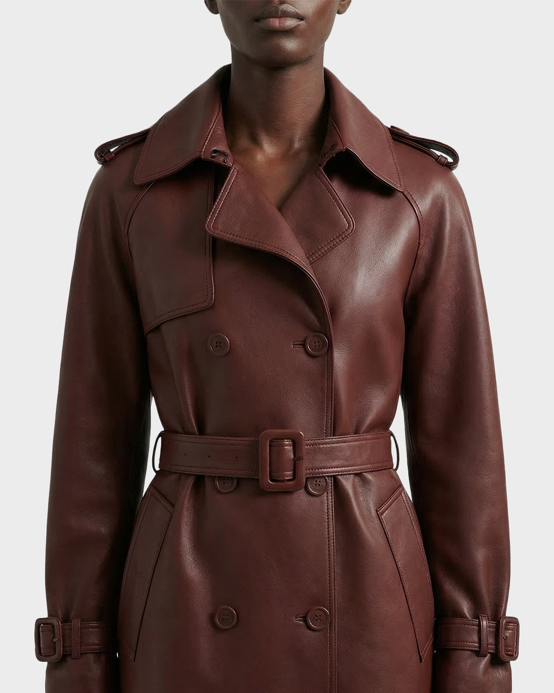 A person wearing the Rudsak CASSIE L-B - 8125553 Rust women's bonded leather long trench coat, featuring a double-breasted silhouette, belted waist, wide lapels, shoulder epaulets, and buttoned cuffs, shown from shoulders to mid-thigh.