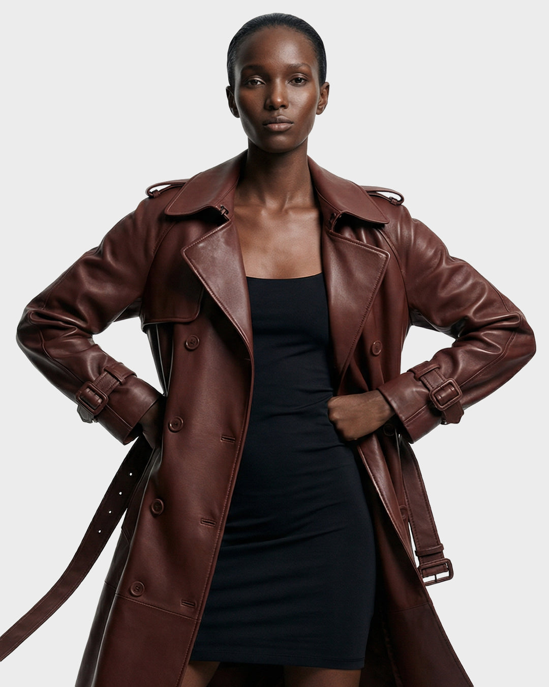 A woman stands confidently with hands on hips, wearing the Rudsak CASSIE L-B - 8125553 Rust long trench coat over a fitted black dress. The double-breasted silhouette contrasts with the plain, light-toned background.