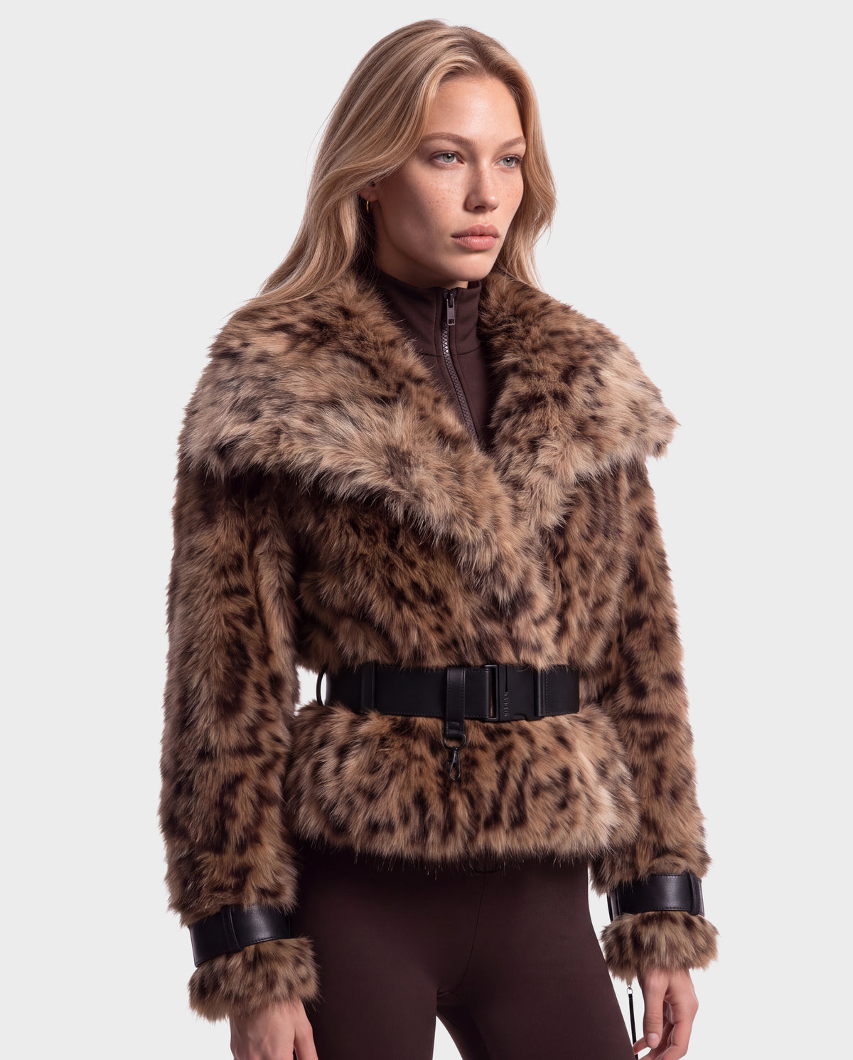 A woman wears the Rudsak CHLOE - 8125619 Leopard, a leopard-print cropped faux fur jacket with a wide black belt at the waist and vegan leather trim on the cuffs, perfect for a winter wardrobe.
