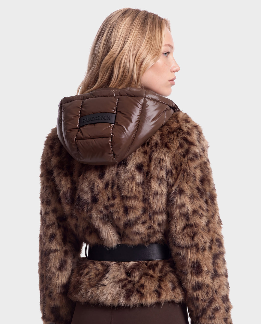 A person wearing a Rudsak CHLOE - 8125619 Leopard faux fur jacket with a brown puffer hood and a black waist belt.