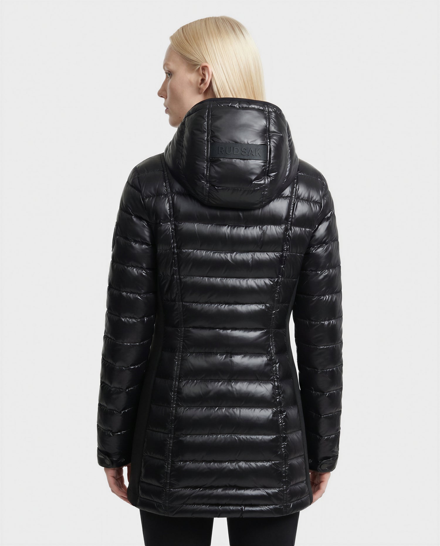 A person with straight blonde hair is seen from behind wearing the Rudsak COSETTE C - L124011 Black, a shiny black quilted hooded jacket. "RUDSAK" is visible on the back of the hood against a plain white background.