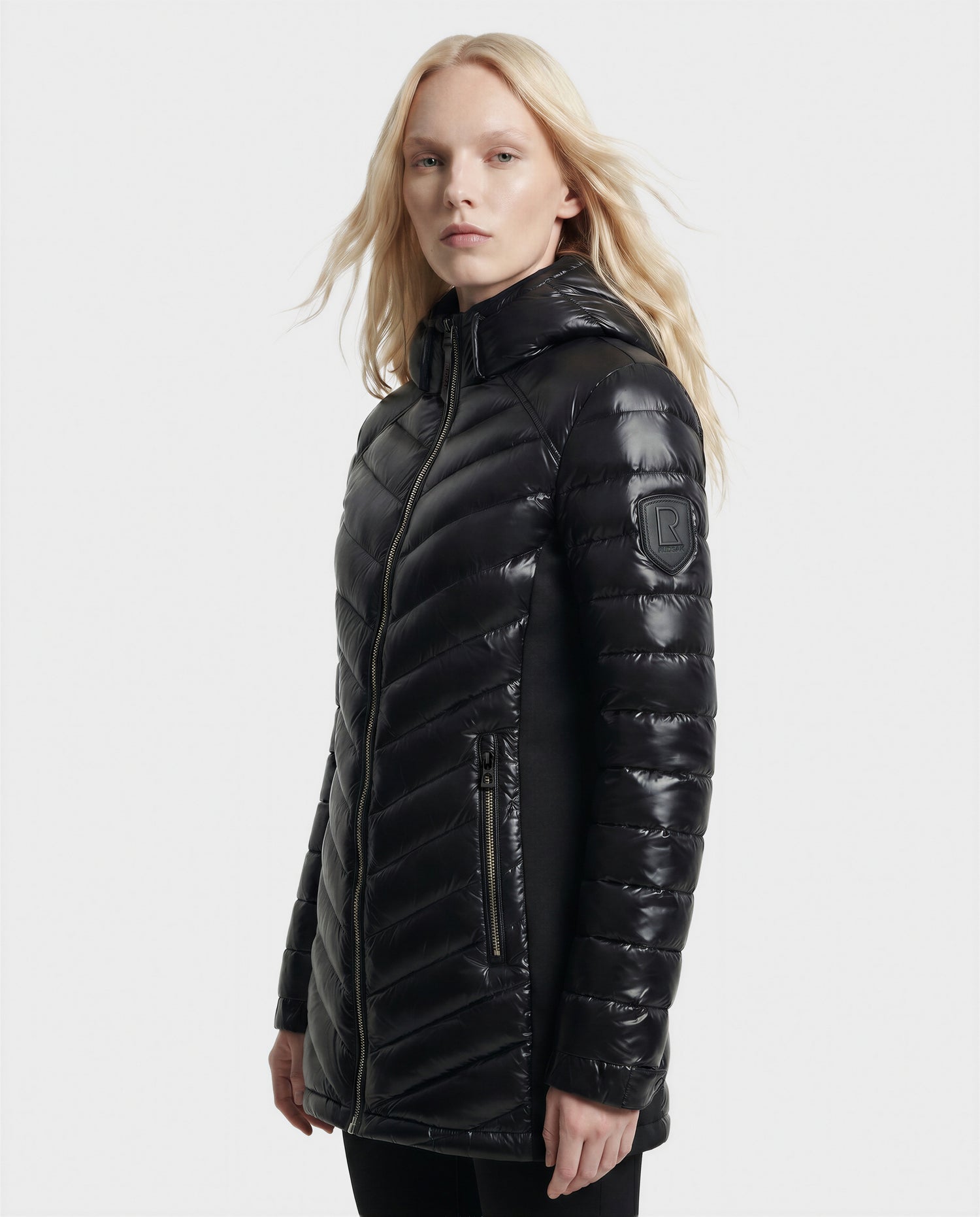 A person with long blonde hair wears the Rudsak COSETTE C - L124011 Black, a glossy black quilted jacket with a hood and zippered pockets, standing against a plain light background.