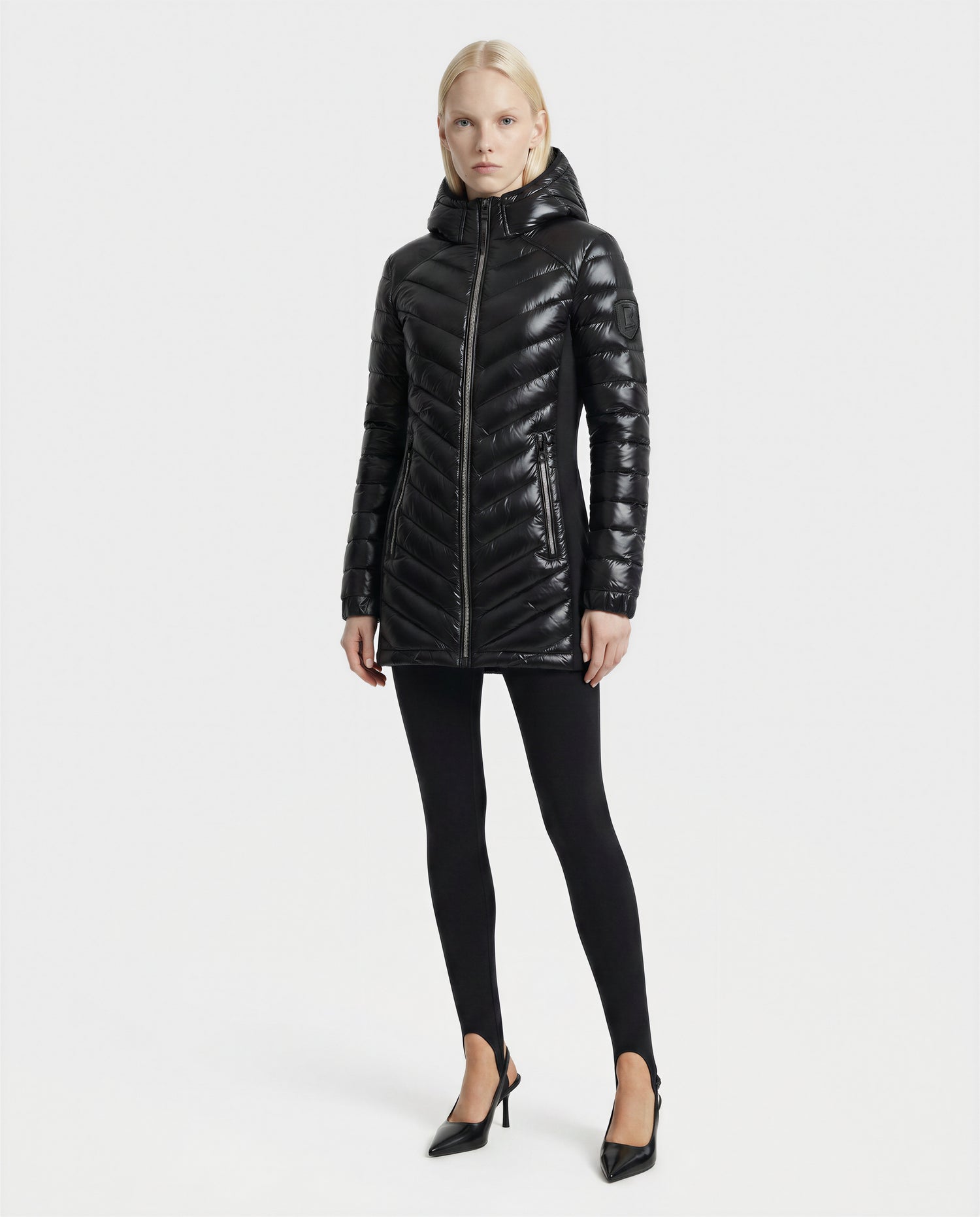 A woman faces forward against a plain white background, wearing the Rudsak COSETTE C - L124011 Black glossy hooded puffer coat, paired with black leggings and black heels.