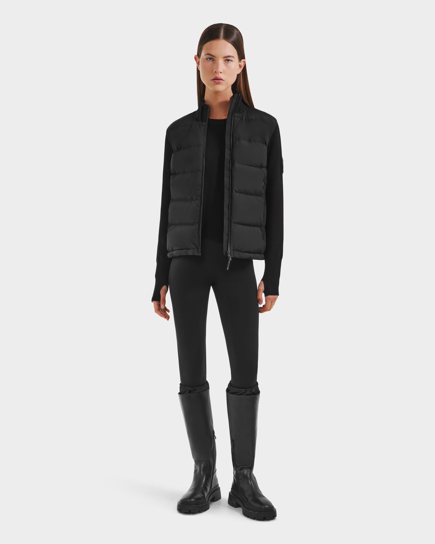 A woman faces forward against a white background, wearing the Rudsak CAMILLE HV - H125500 Black limited-edition down jacket over a black long-sleeve top, black leggings, and tall black boots.