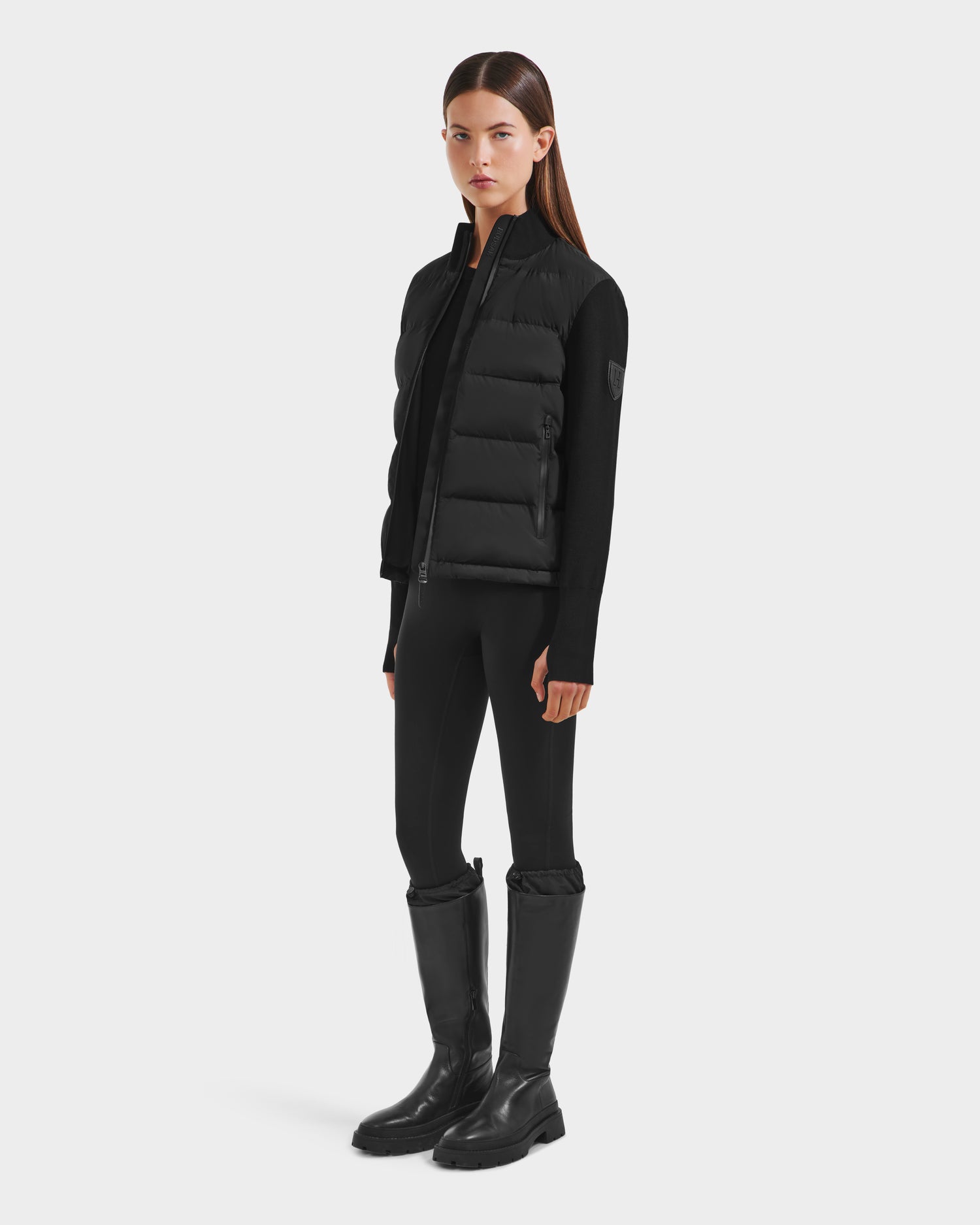 A woman faces forward against a white background, wearing Rudsak’s CAMILLE HV - H125500 Black limited-edition down jacket over a black long-sleeve top, fitted black pants, and knee-high black boots.