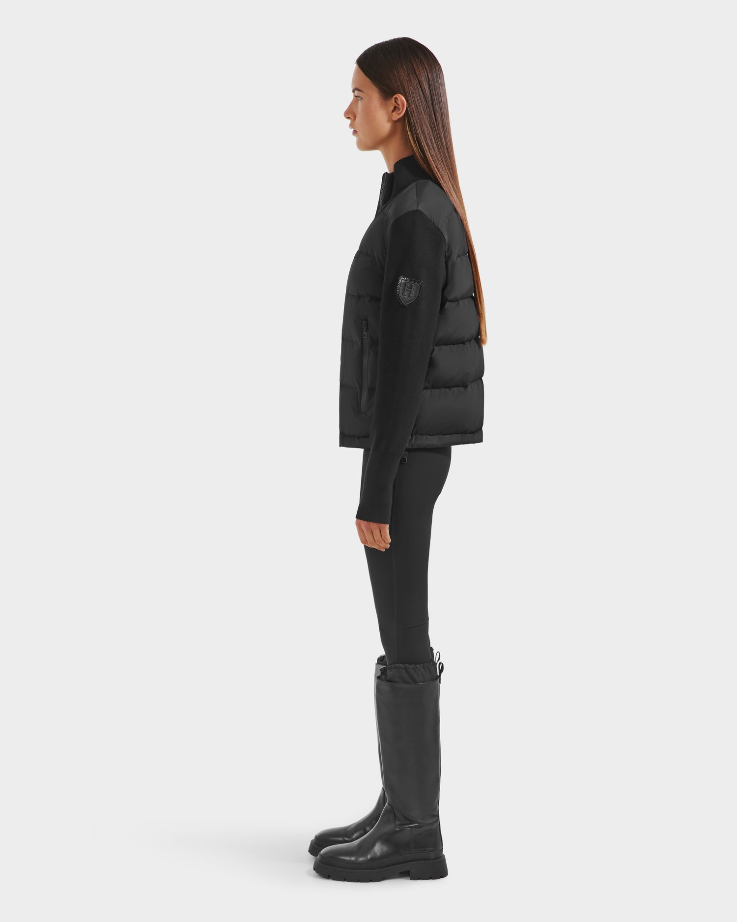 A woman in profile stands against a white background, wearing the Rudsak CAMILLE HV - H125500 Black jacket, black leggings, and tall black boots. Her straight brown hair is loose as she looks forward.