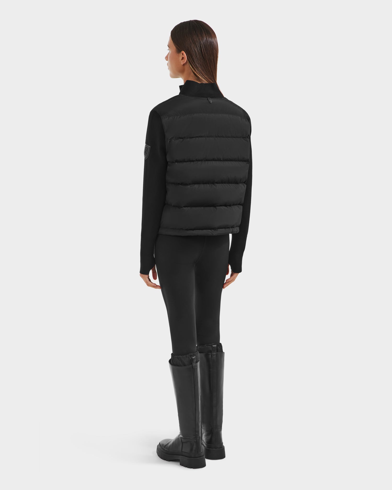 A woman with straight brown hair faces away, wearing the limited-edition Rudsak CAMILLE HV - H125500 Black down jacket over a black long-sleeve top, black leggings, and tall black boots against a plain white background.