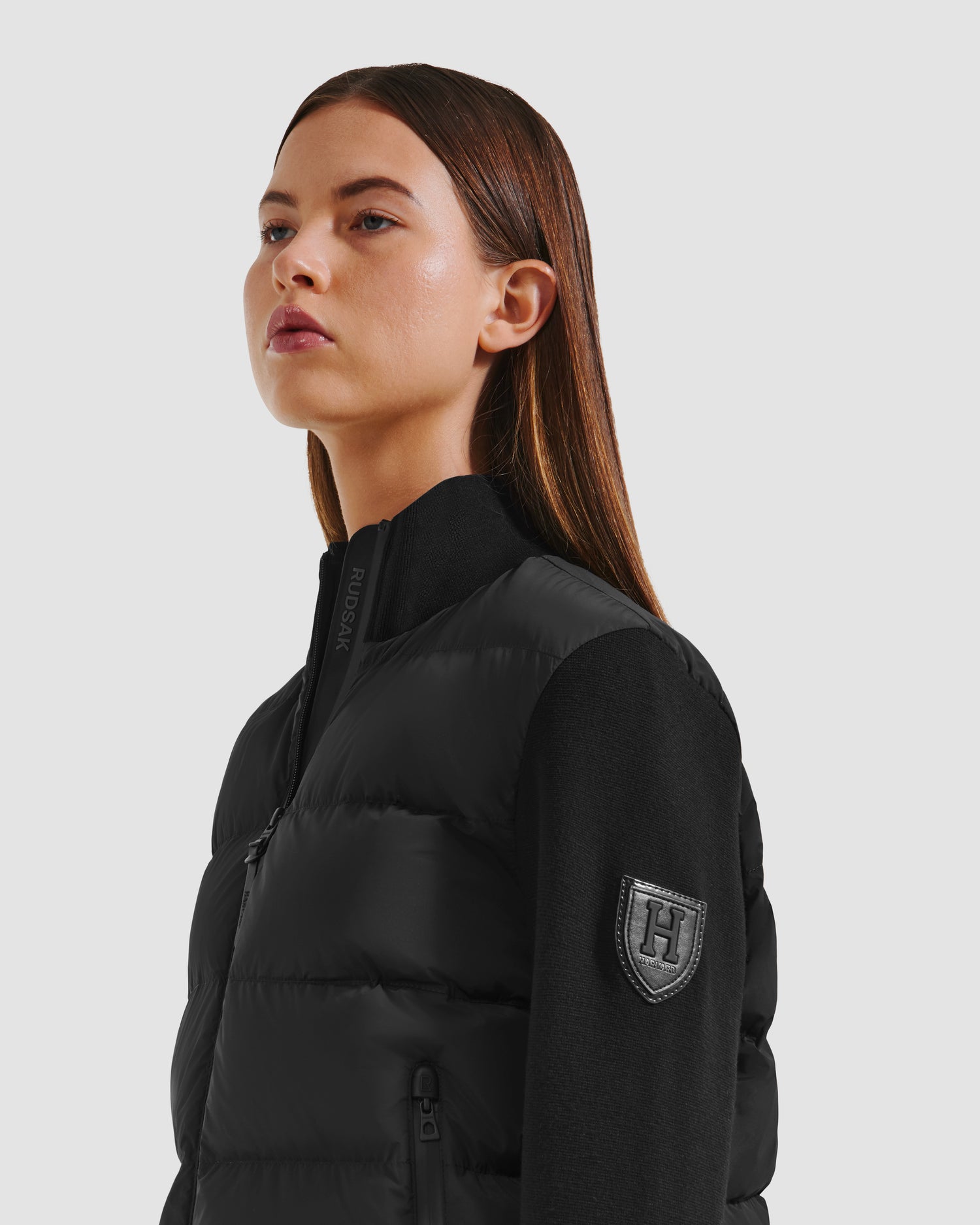 A woman with straight brown hair wears the Rudsak CAMILLE HV - H125500 Black down jacket, featuring a zipper and an H patch on the sleeve, standing against a plain light background.