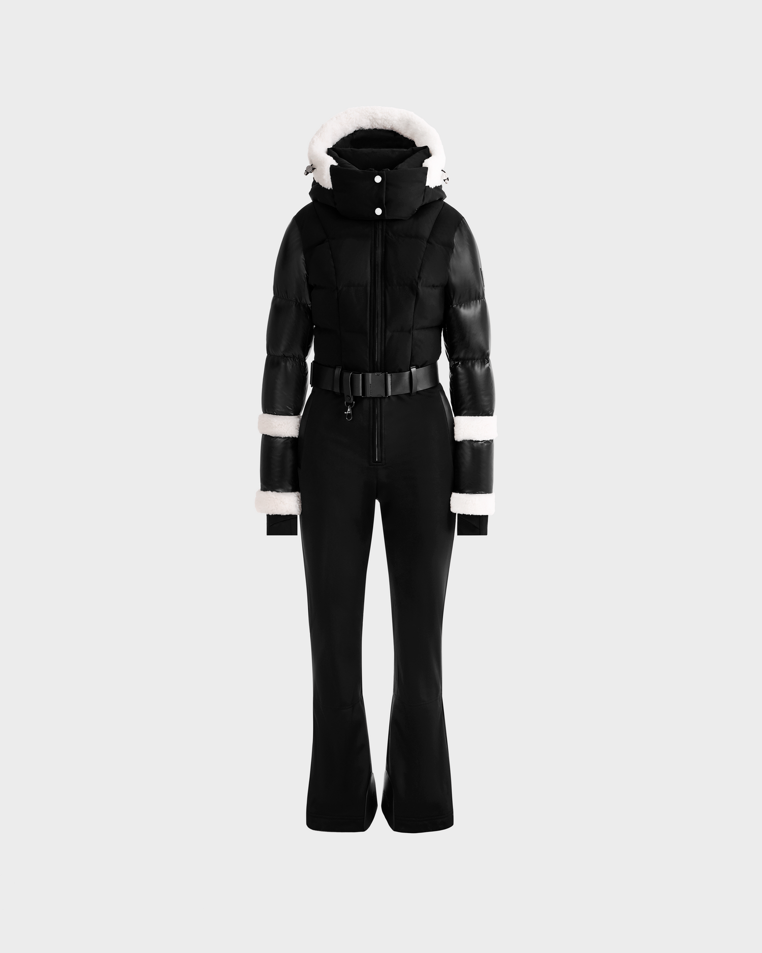 The Rudsak CARMEN - 8125708 Black is a stylish one-piece ski suit with a fitted jacket, white fur trim on the hood, cuffs, and upper arms, matching black flared pants, and a belted waist, shown against a grey background.