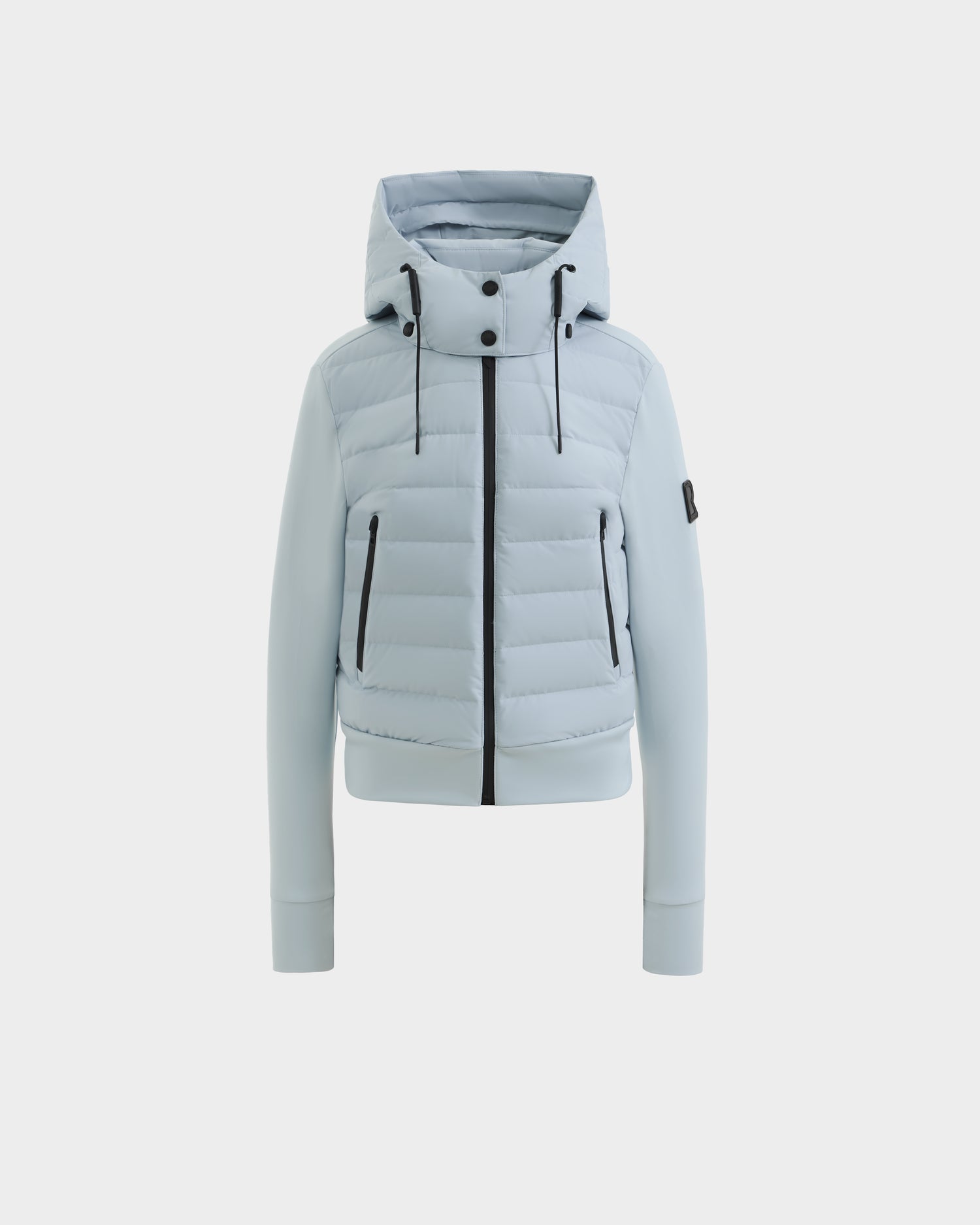 The Rudsak CASSA - 8125057 Powder Blue women's jacket features a hood, front zipper, snap buttons, two zippered side pockets, and a logo patch on the left sleeve, shown against a plain white background.