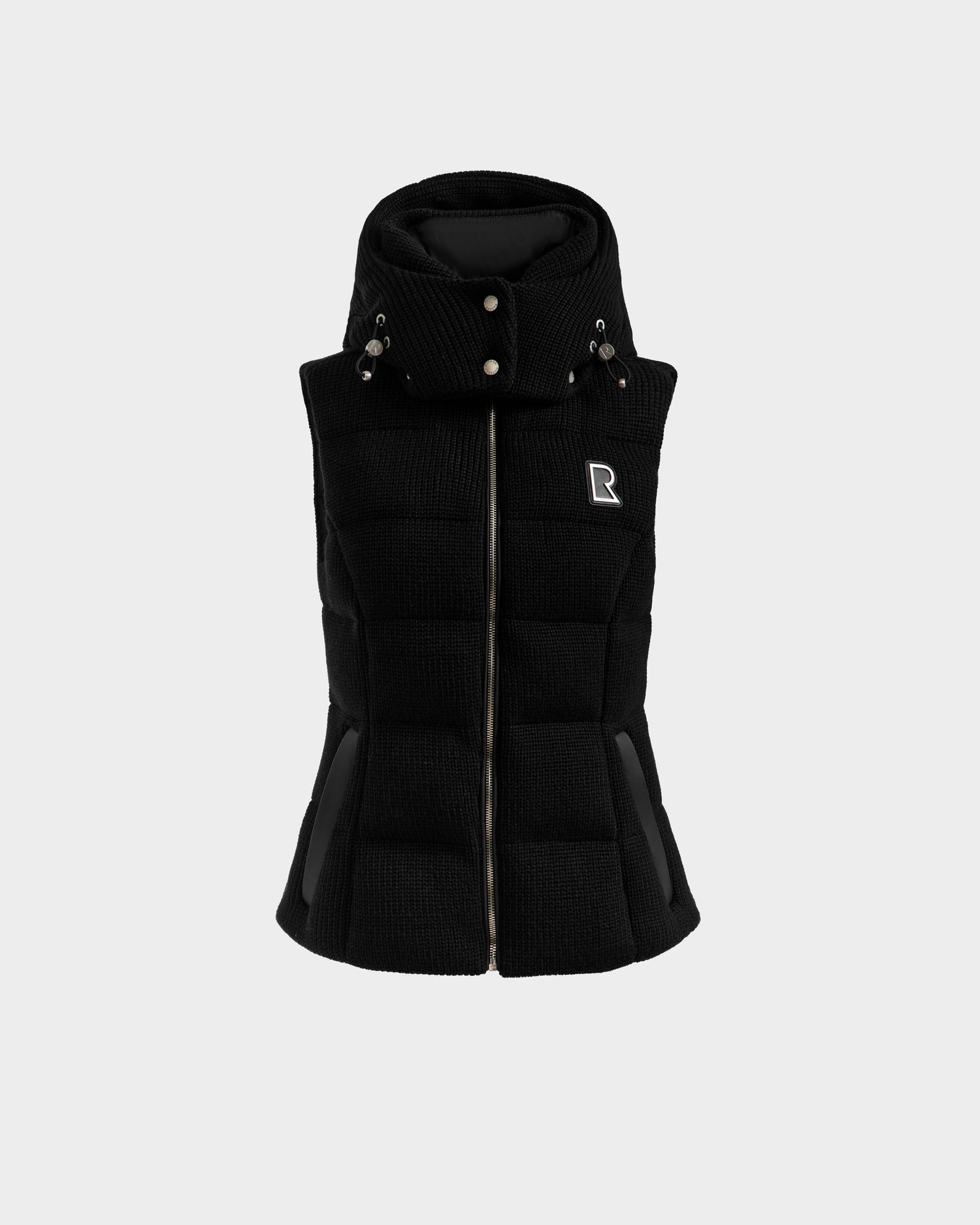 The CASSIDY - 8125622 Black by Rudsak is a black, sleeveless women’s down vest with a high collar, snap buttons, front zipper, side pockets, and an R logo on the chest—ideal for lightweight warmth in transitional weather. Shown on light gray.
