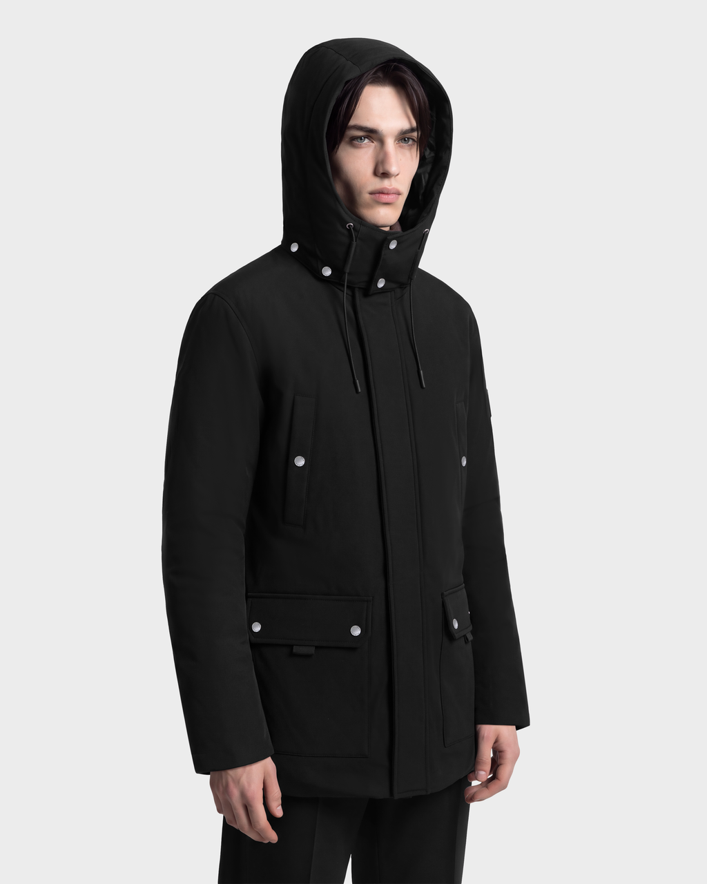 A man wearing a black Rudsak EDWARD - 6126501 Black hooded duck down parka with silver snap buttons and front pockets.