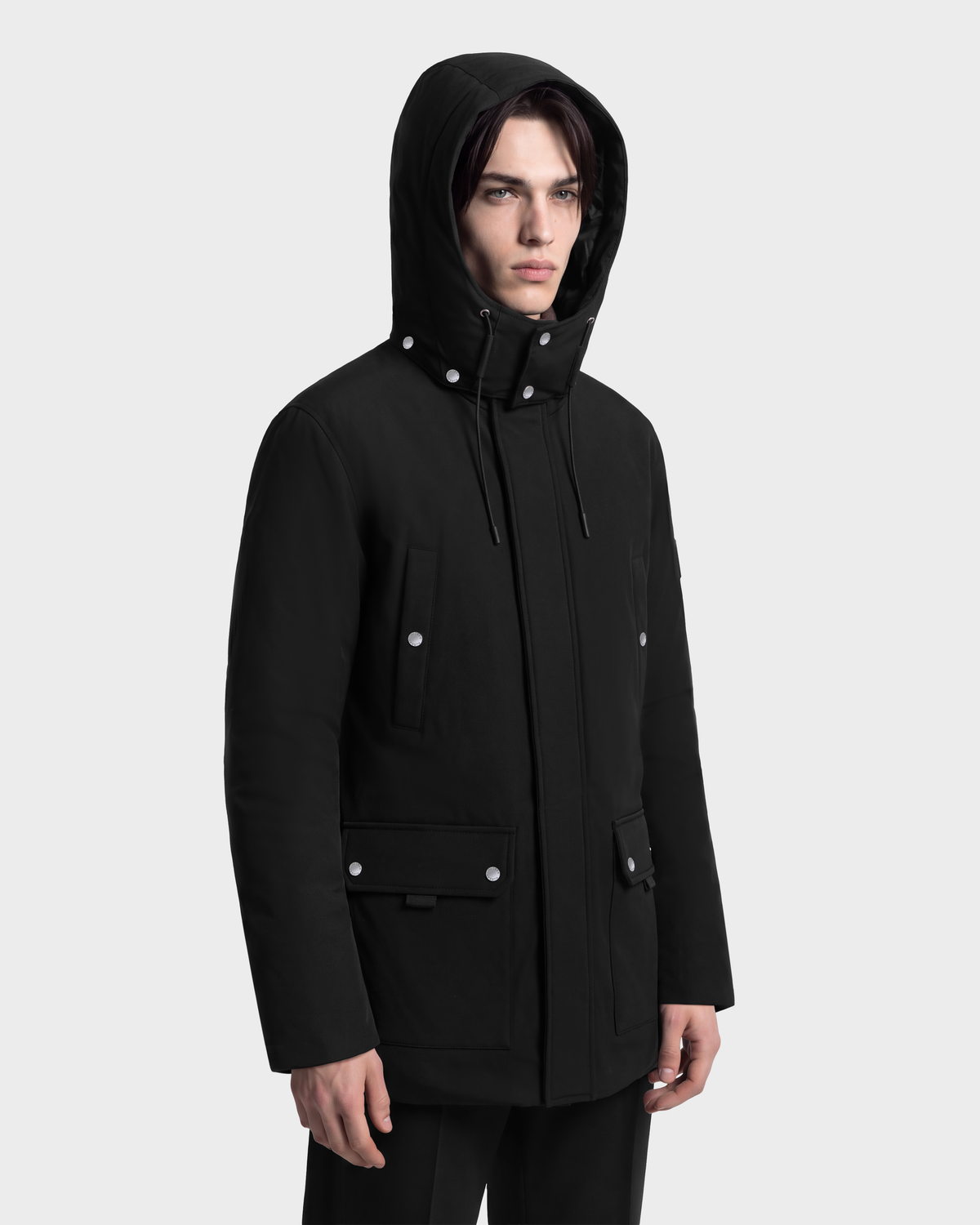 A man wearing a black Rudsak EDWARD - 6126501 Black hooded duck down parka with silver snap buttons and front pockets.