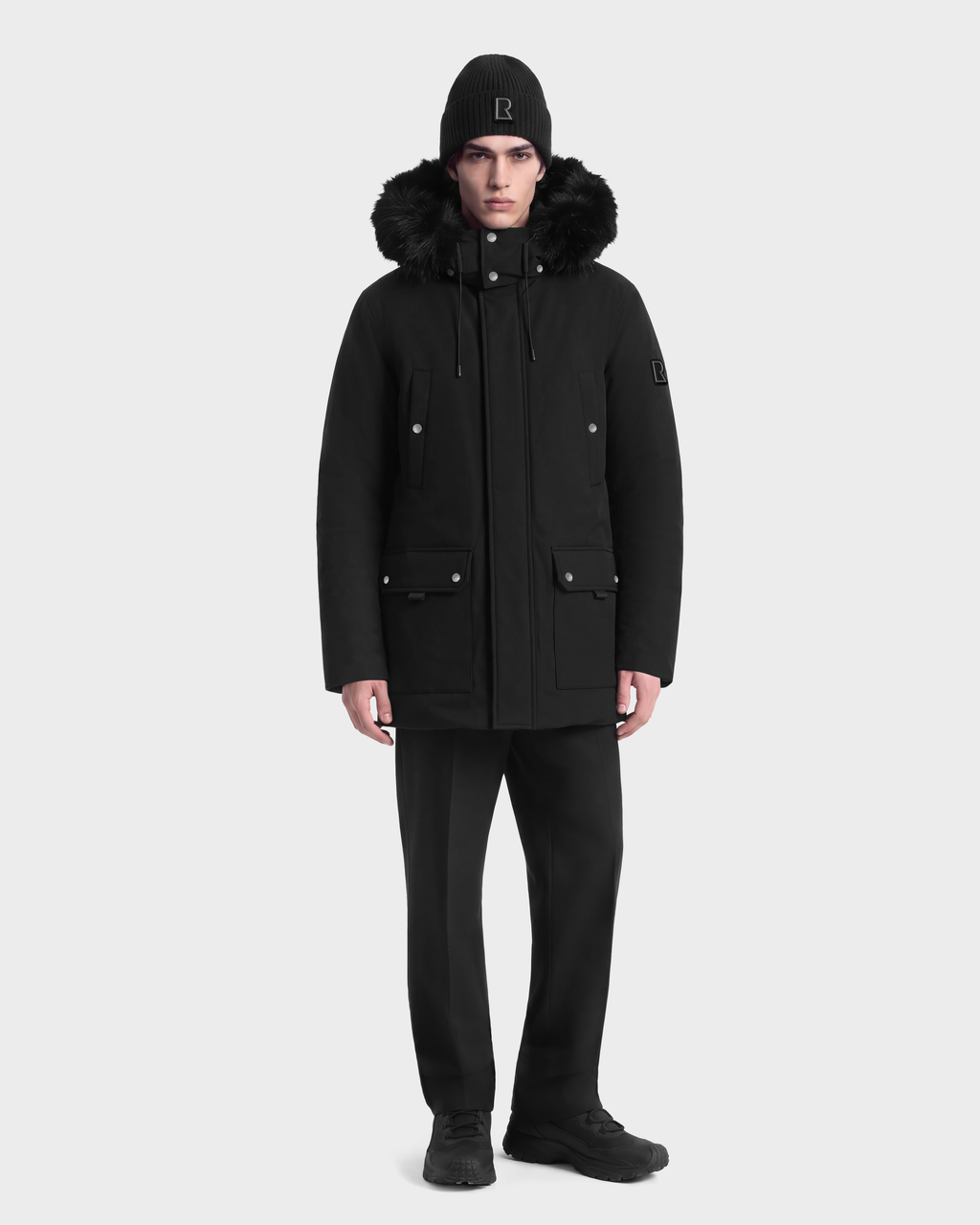 A man wearing an EDWARD - 6126501 Black duck down winter parka with a fur-trimmed hood, black trousers, and a black knit beanie.