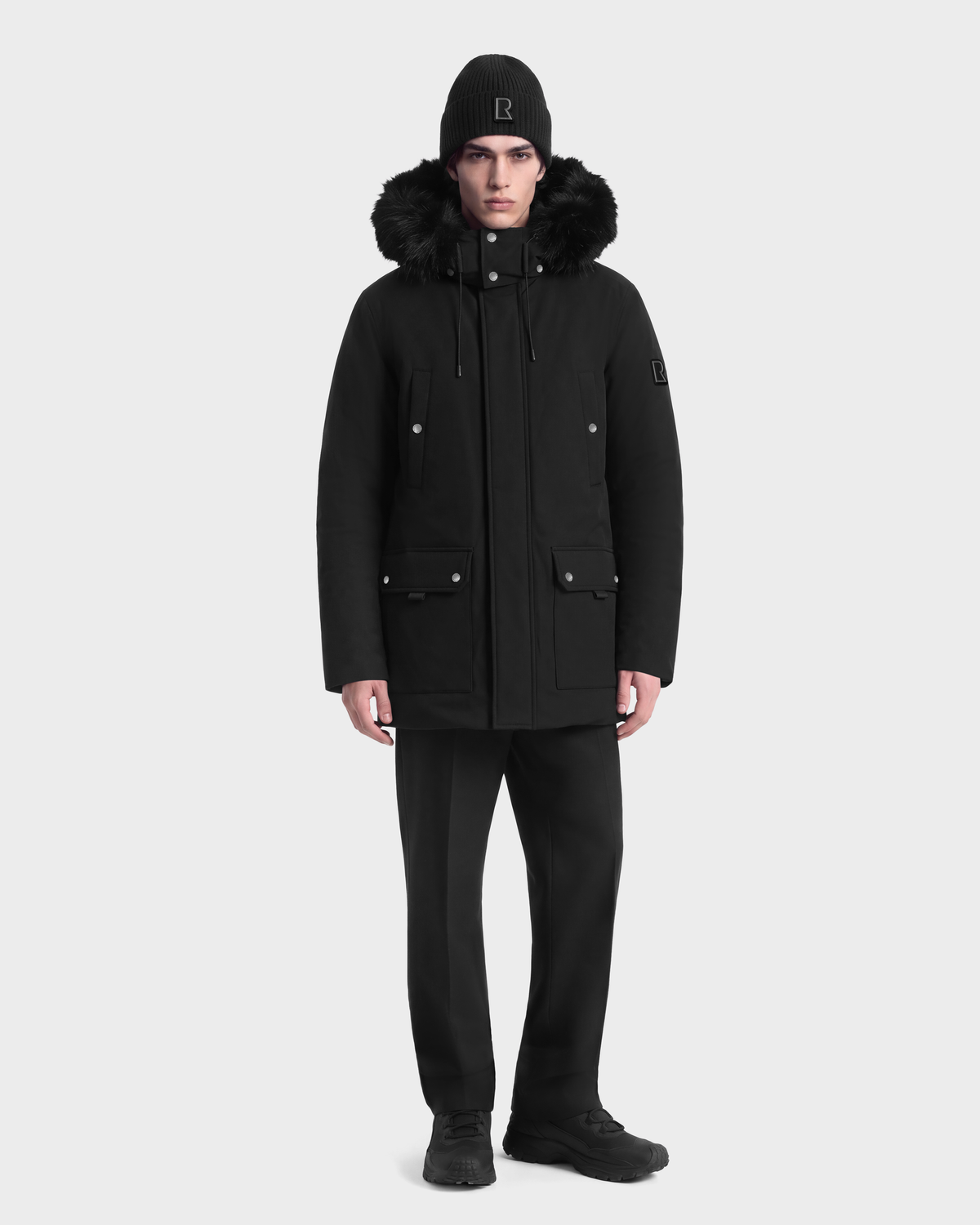 A man wearing an EDWARD - 6126501 Black duck down winter parka with a fur-trimmed hood, black trousers, and a black knit beanie.