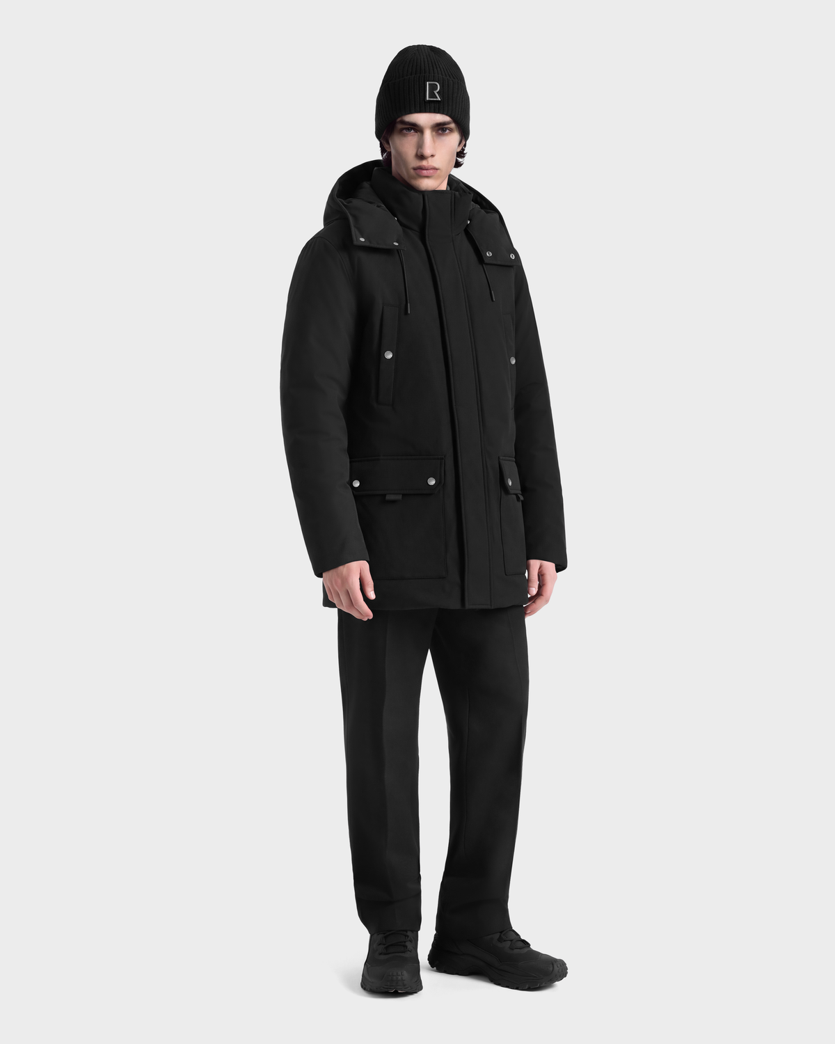 A person wearing a black EDWARD - 6126501 Black hooded winter parka, black trousers, and a black beanie with a logo on the front.