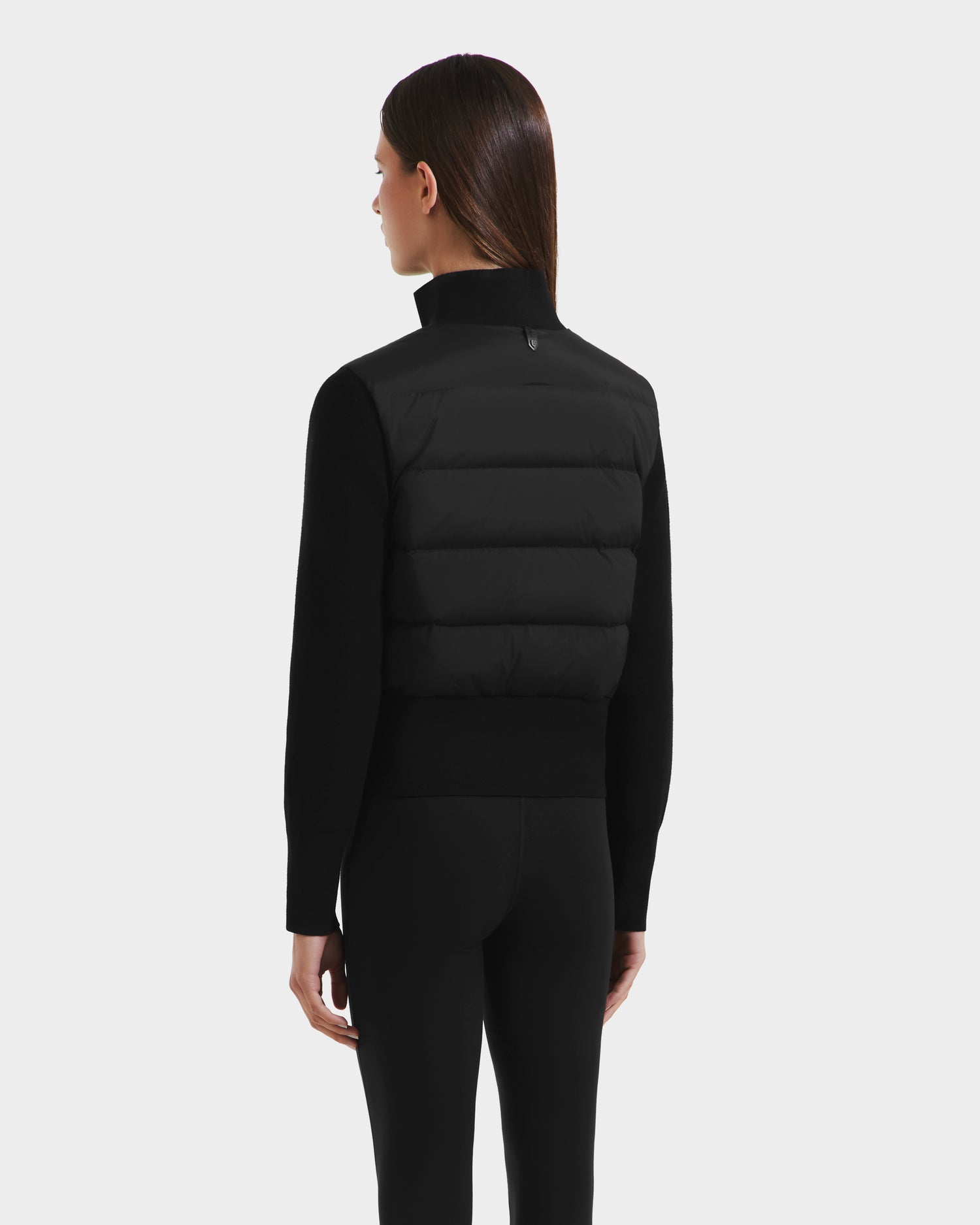 A woman with straight brown hair is shown from the back, wearing the Rudsak CLAREMONT - 8125502 Black RDS-certified down padded jacket with a high collar and fitted black pants against a plain light gray background.