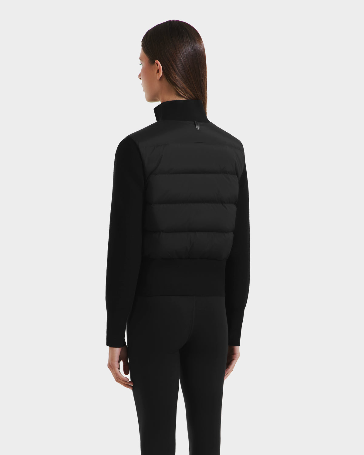 A woman with straight brown hair is shown from the back, wearing the Rudsak CLAREMONT - 8125502 Black RDS-certified down padded jacket with a high collar and fitted black pants against a plain light gray background.