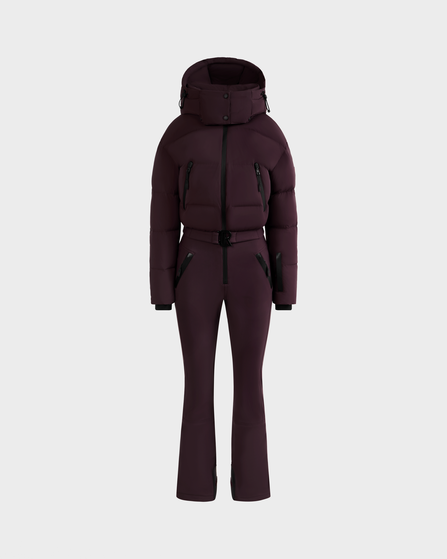 The DINARA - 8125600 Bordeaux by Rudsak is a dark purple, fitted women’s one-piece ski suit with a belted waist, puffer-style hooded jacket, black zipper details, and long sleeves, shown on a plain light gray background.