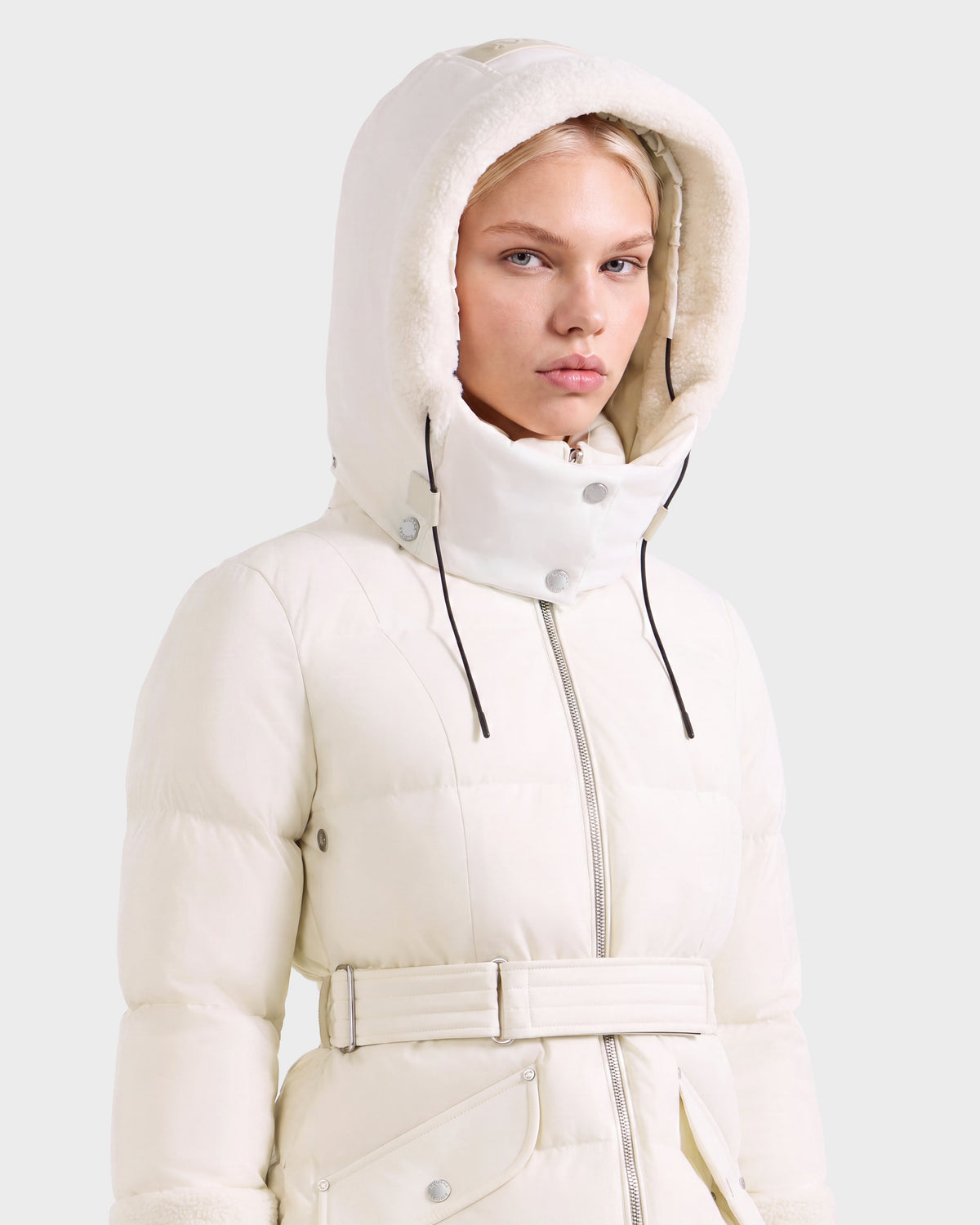 A person wearing a Rudsak AUDREY L - 8124585 Vanilla knee-length down puffer jacket with a belted waist and a large faux shearling-lined hood.
